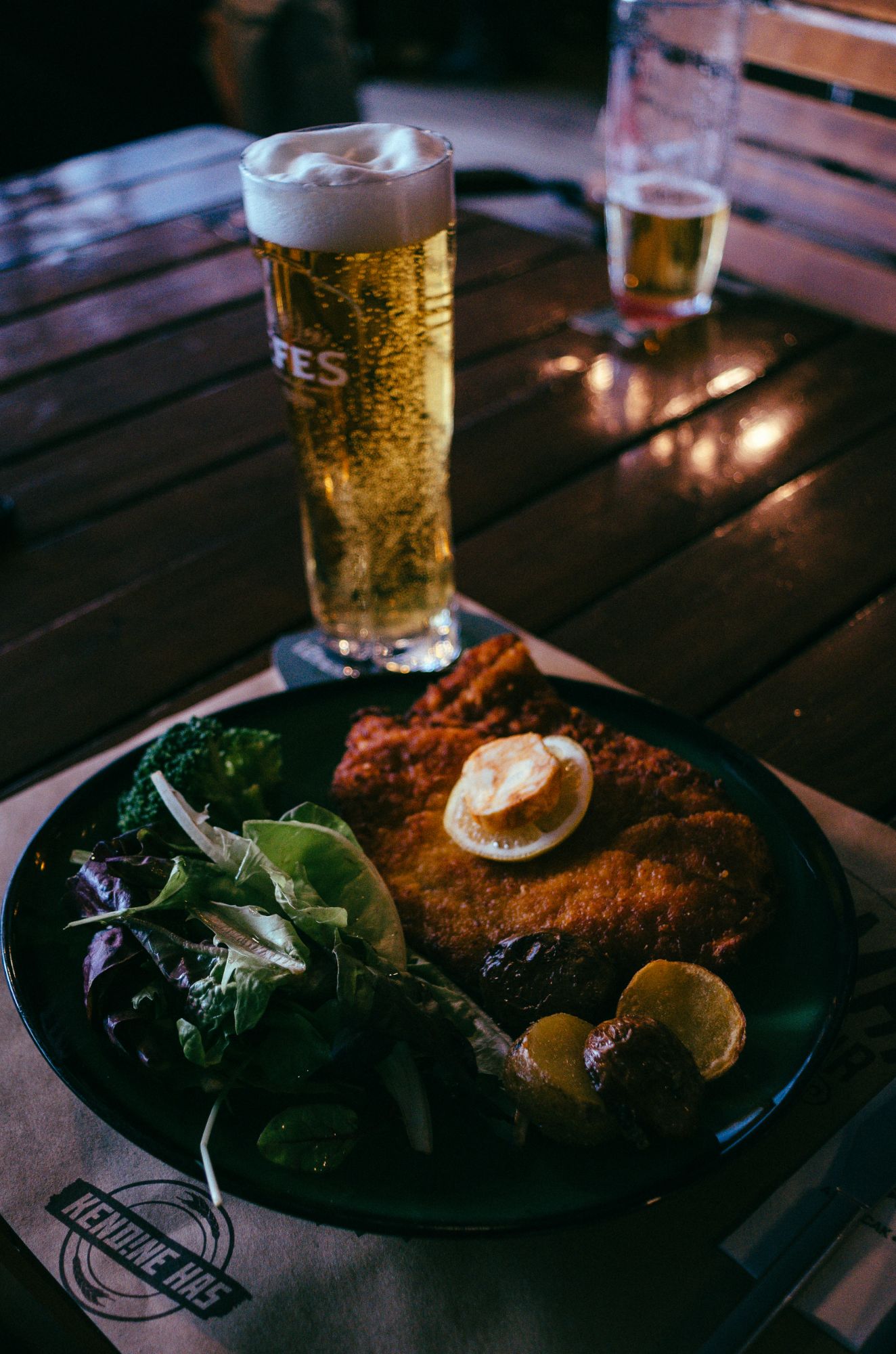 Biere blondă și un platou cu carne pane, legume proaspete și felii de lămâie, servite pe o masă din lemn.