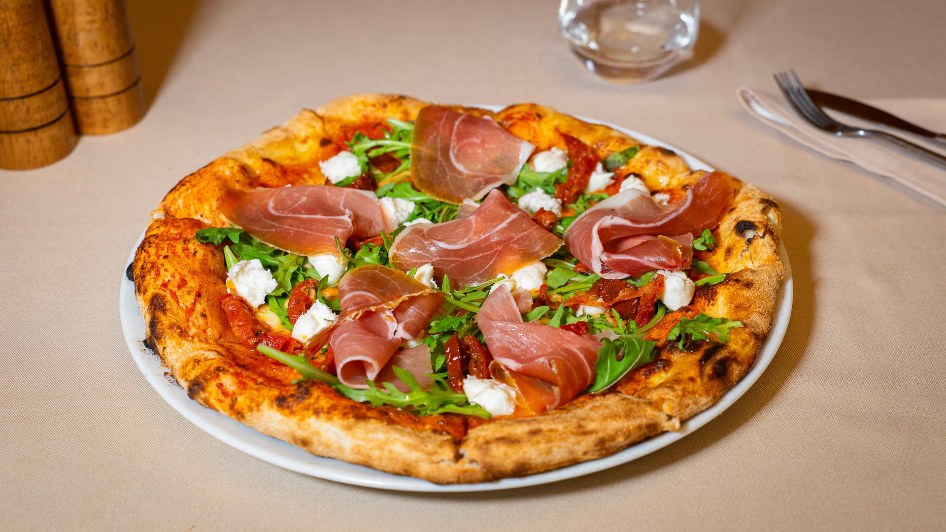 Pizza cu prosciutto, mozzarella, roșii uscate și rucola pe un fundal de masă elegantă.
