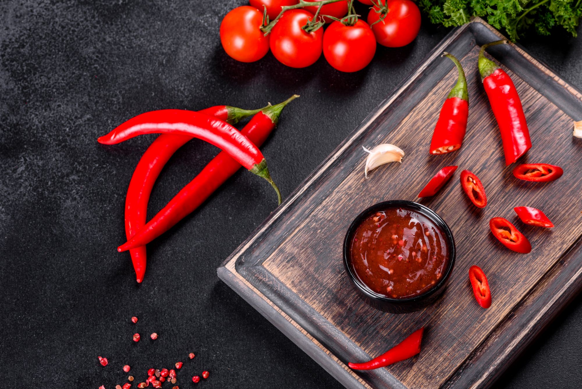 Sos chili w miseczce, świeże papryczki, czosnek i pomidory na drewnianej desce.