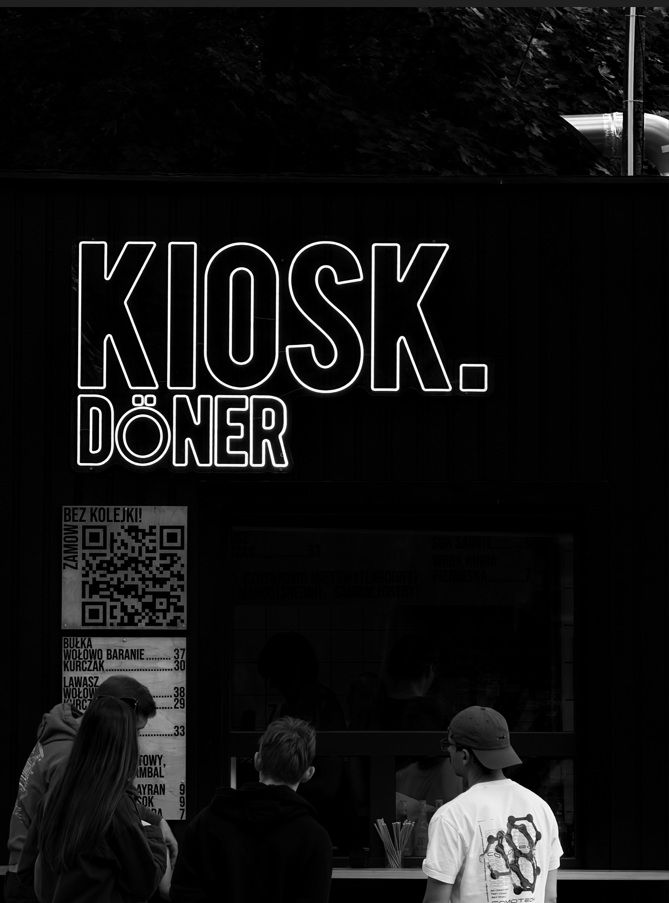 Kiosk z jedzeniem, z wyraźnym napisem "KIOSK. DÖNER" na tle czarnej ściany, klienci w kolejce.