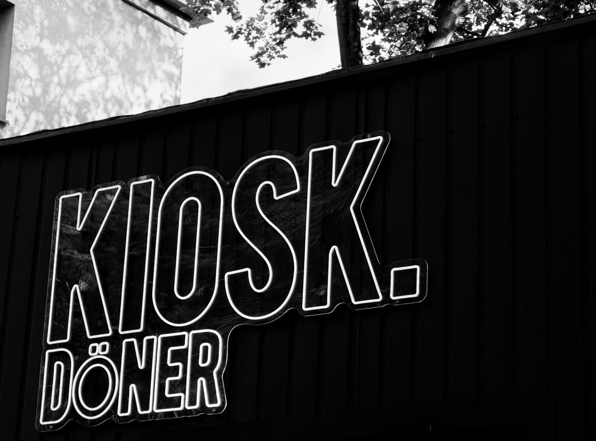 Czarnobiała fotografia pokazująca neonowy szyld "KIOSK. DÖNER" na ścianie budynku.