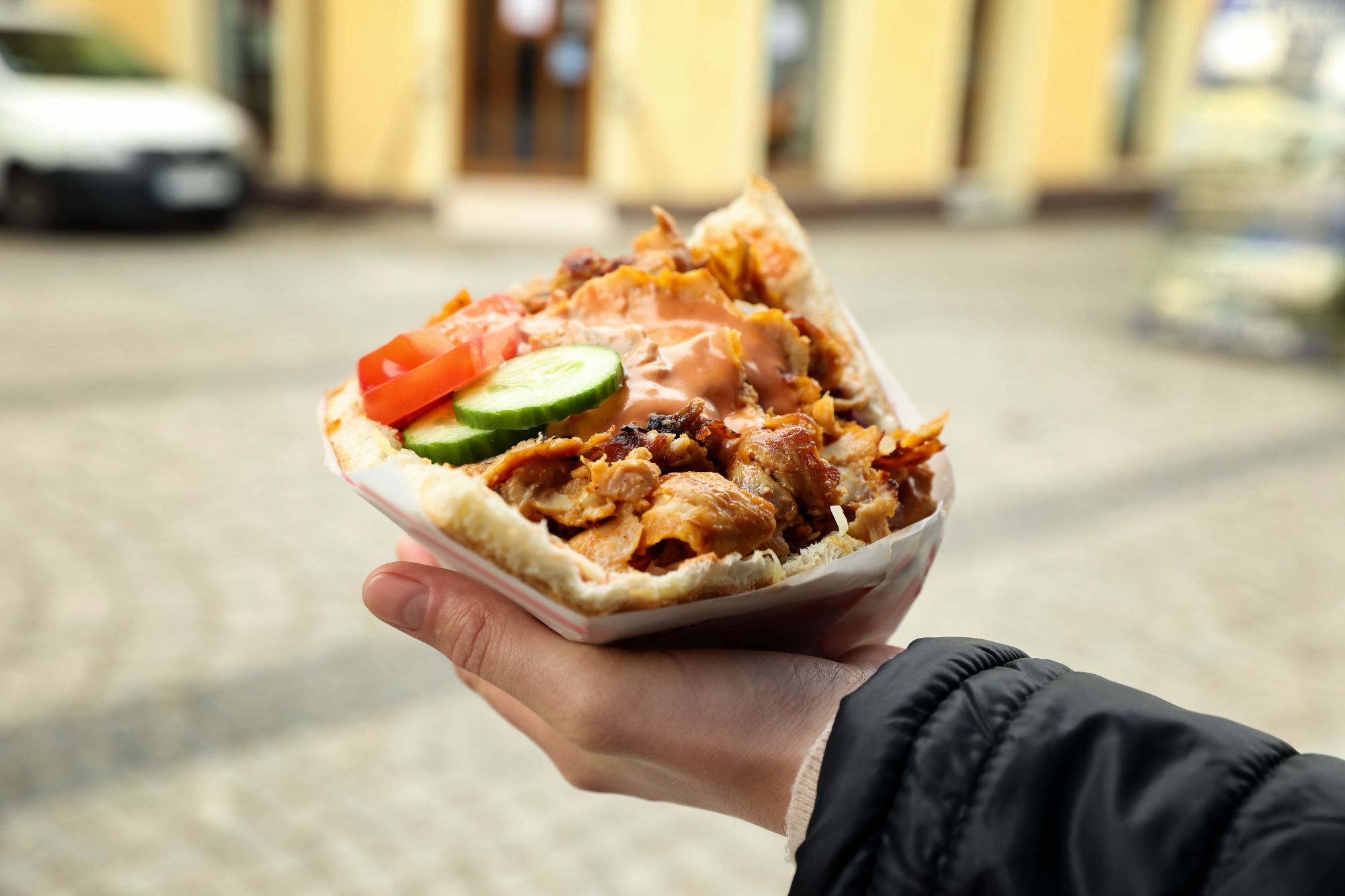 Kanapka z kurczakiem, frytkami, pomidorem i ogórkiem, trzymana w ręku na tle miejskiej ulicy.