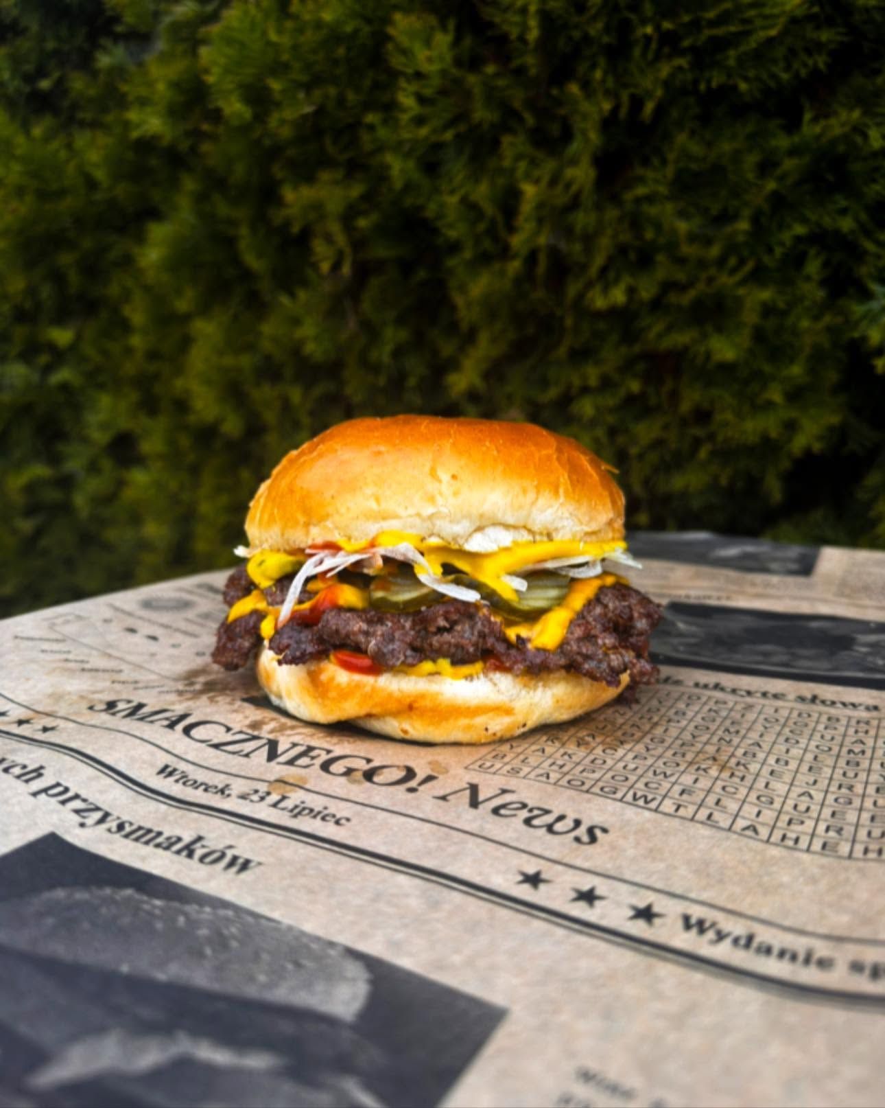 Soczysty hamburger z wołowiną, serem, ogórkiem, cebulą i sałatą na tle gazetowej makiety.