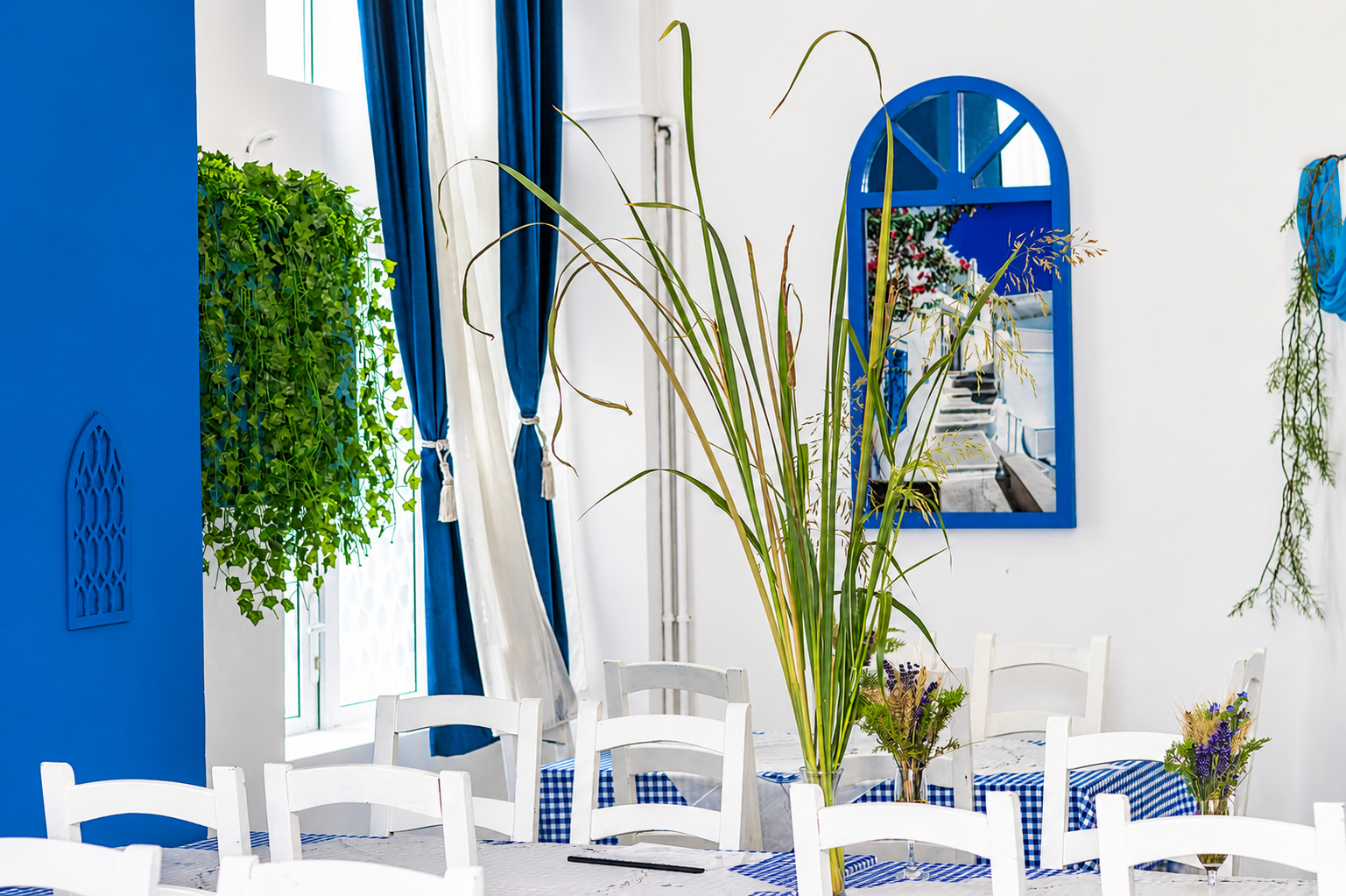 Interior cu mese albe, decor albastru, plante verzi și un oglindă reflectând un peisaj luminos.