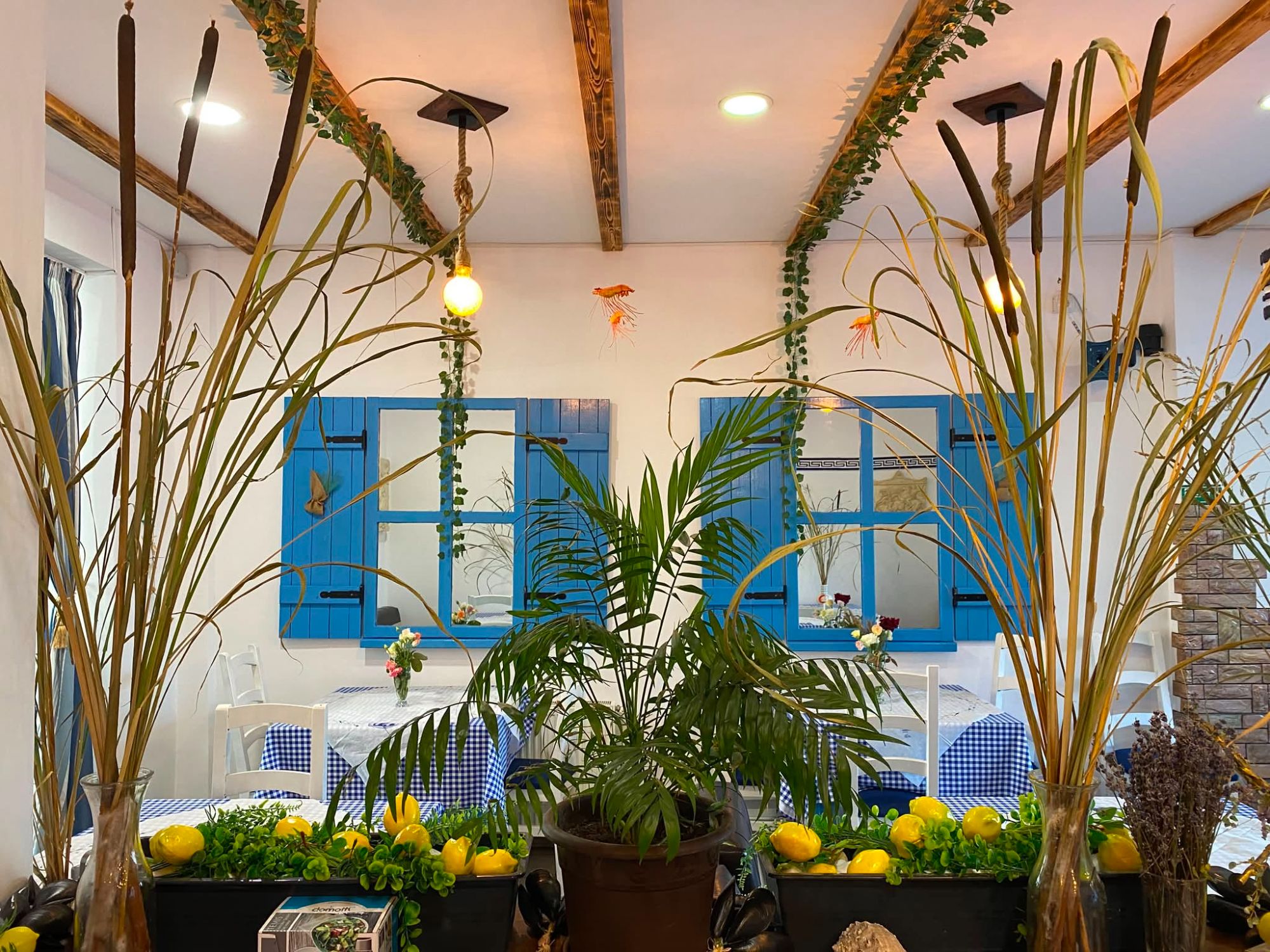 Interior de restaurant cu plante verzi, lămâi, mese albe și feronerie albastră, decor rustic și lumini calde.