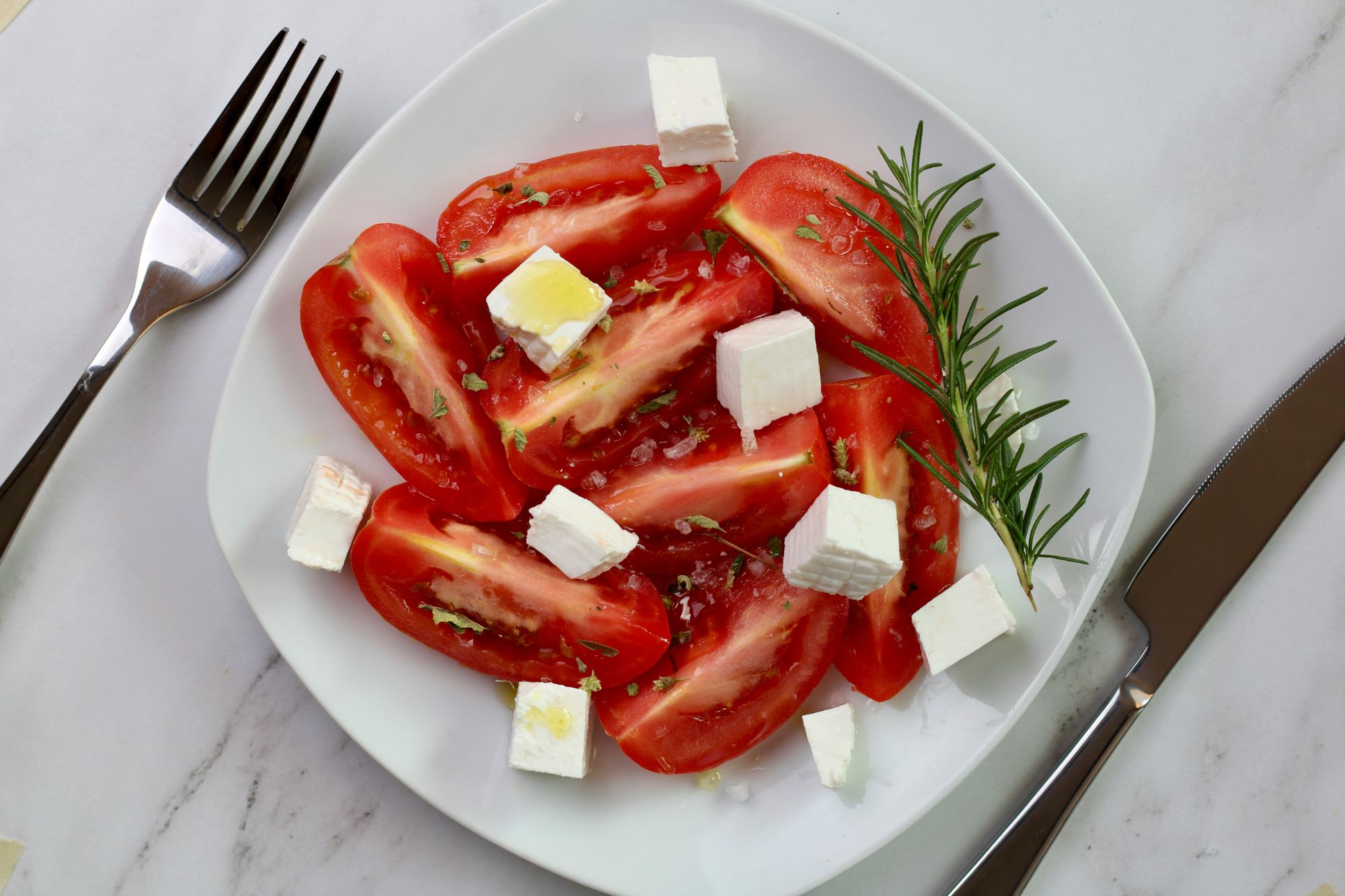 Salată de roșii cu brânză feta, ulei de măsline și rozmarin, servită pe un platou alb.