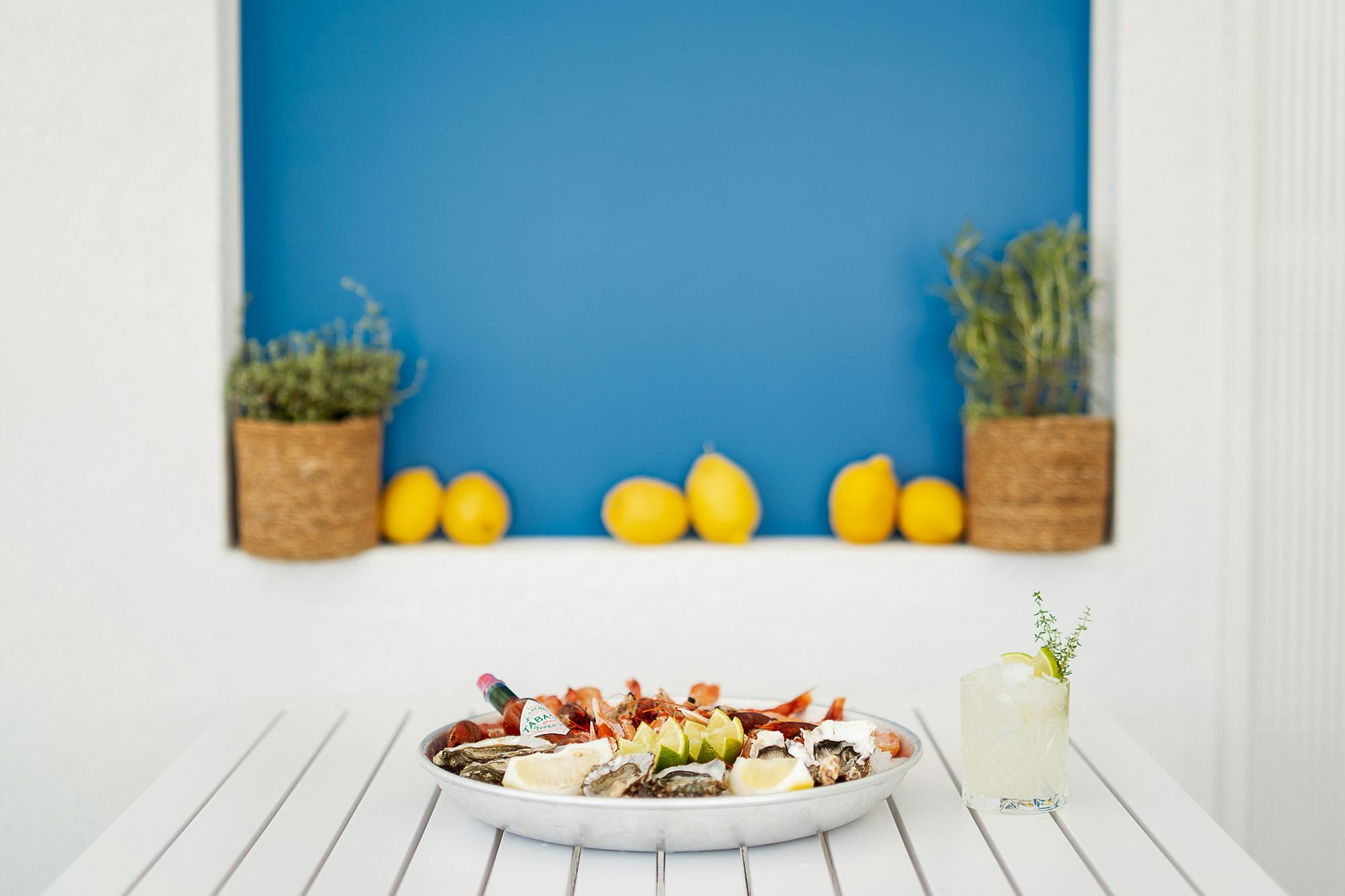 Plată cu fructe de mare, brânzeturi și lămâi pe un fundal albastru. Cocktail cu ierburi pe masă albă.