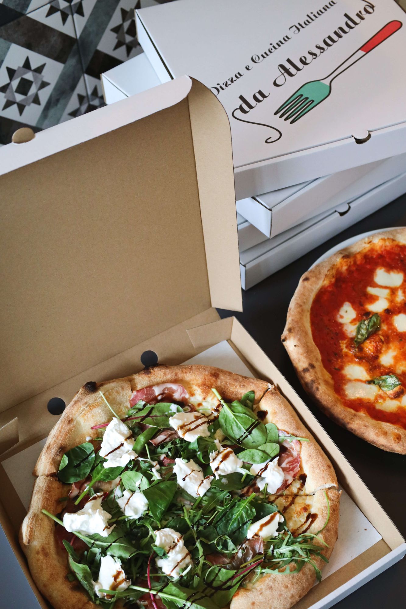 Pizza w pudełku z sałatą, serem i szynką, obok inna pizza margherita w opakowaniu.
