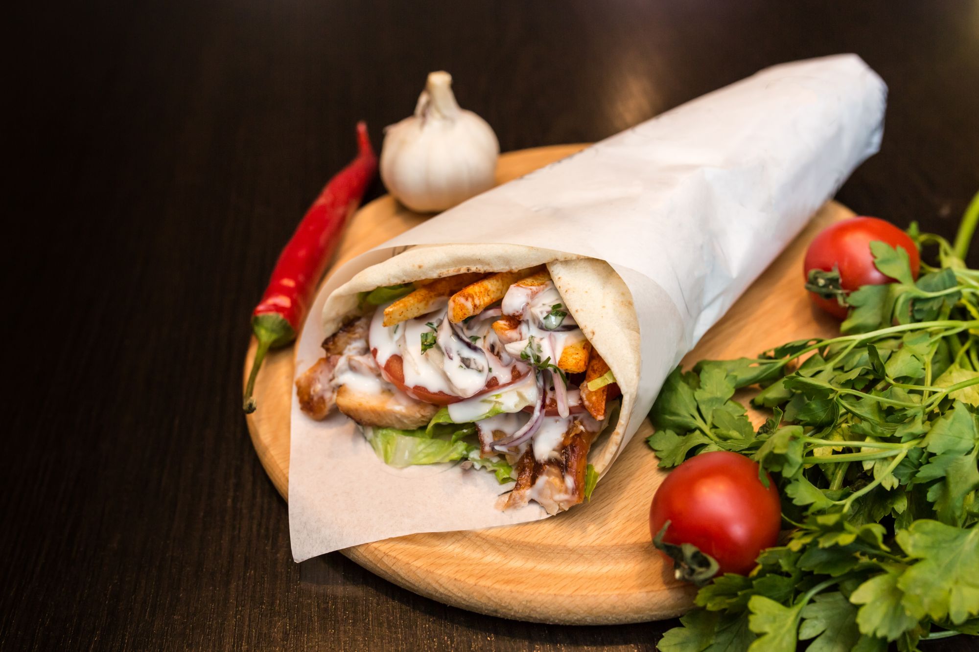 Kebab w papierze, sałata, pomidory, czosnek i kolendra na drewnianej desce.