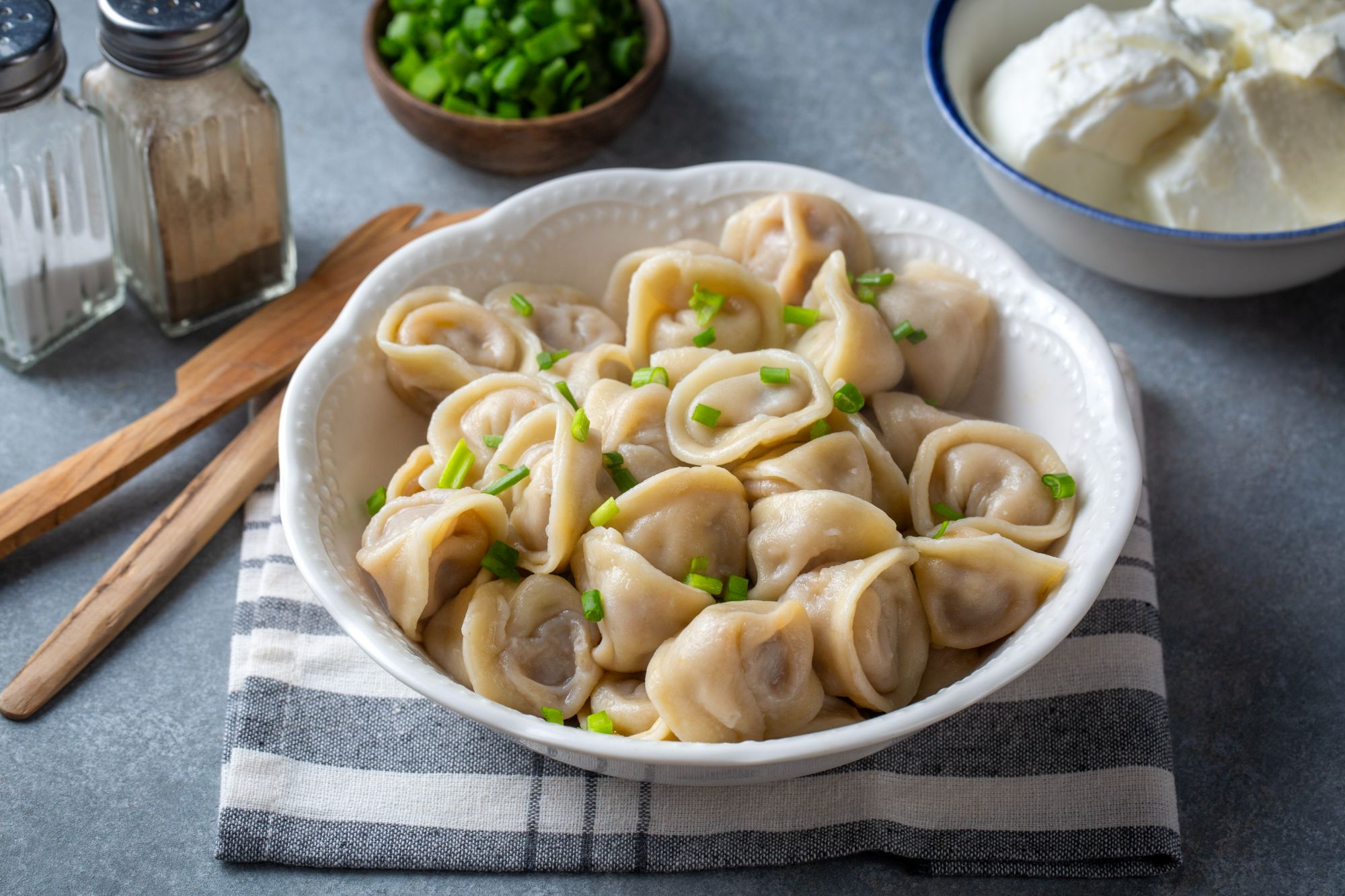 Pierogi z zieloną cebulką w białej misce, obok sól, pieprz, śmietana i drewniane sztućce.
