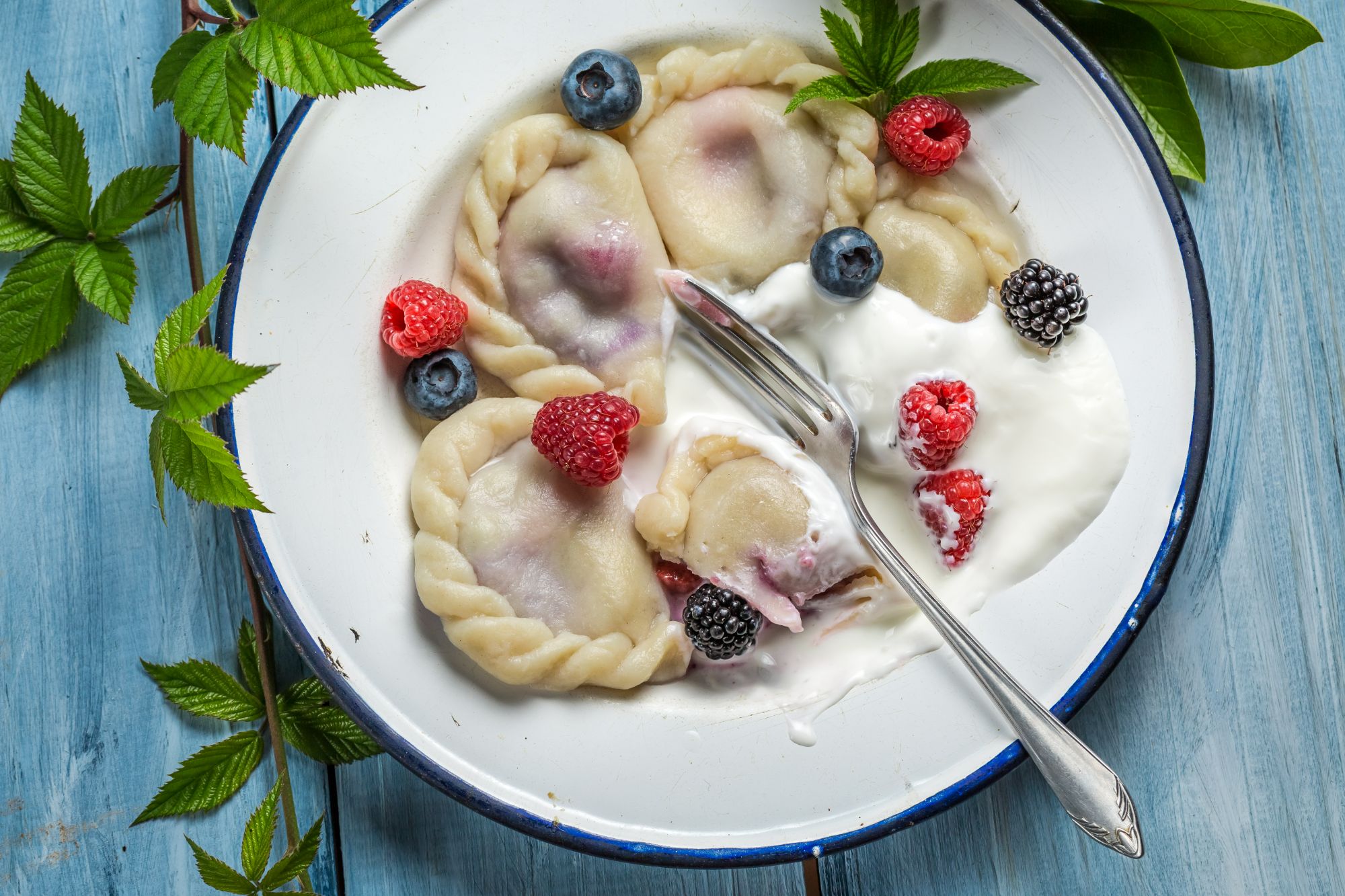 Pierogi z owocami, malinami, borówkami, podane z jogurtem w miseczce na drewnianym tle.