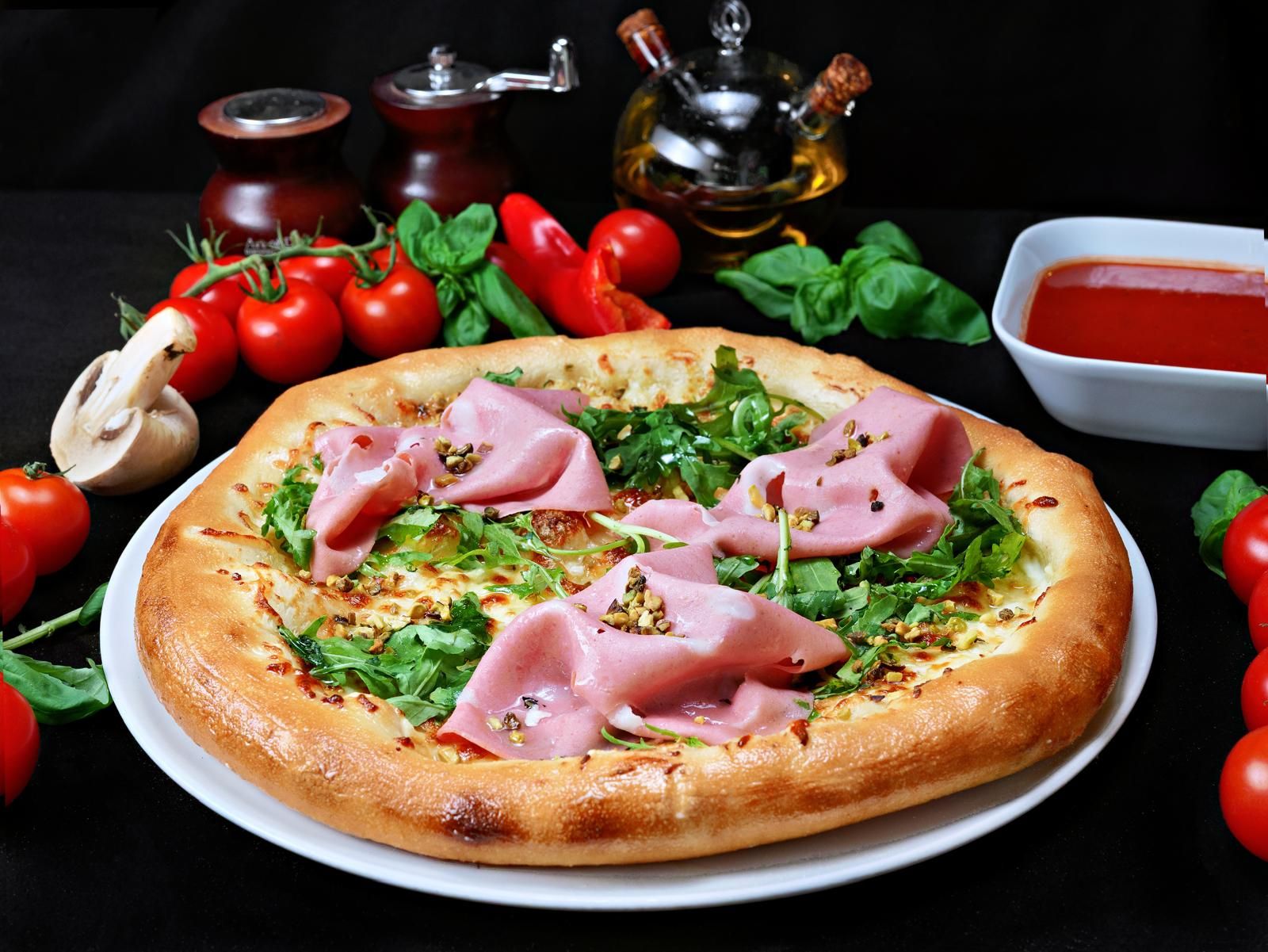 Pizza cu șuncă și salată, înconjurată de roșii proaspete și ingrediente culinare pe un fundal negru.