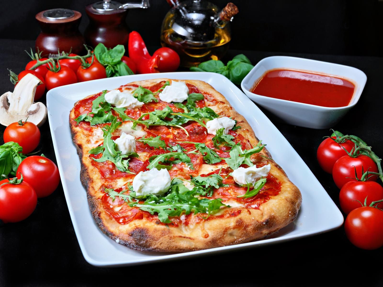 Pizza cu sos de roșii, mozzarella, salam picant, rucola și roșii proaspete, servită pe un fundal negru.