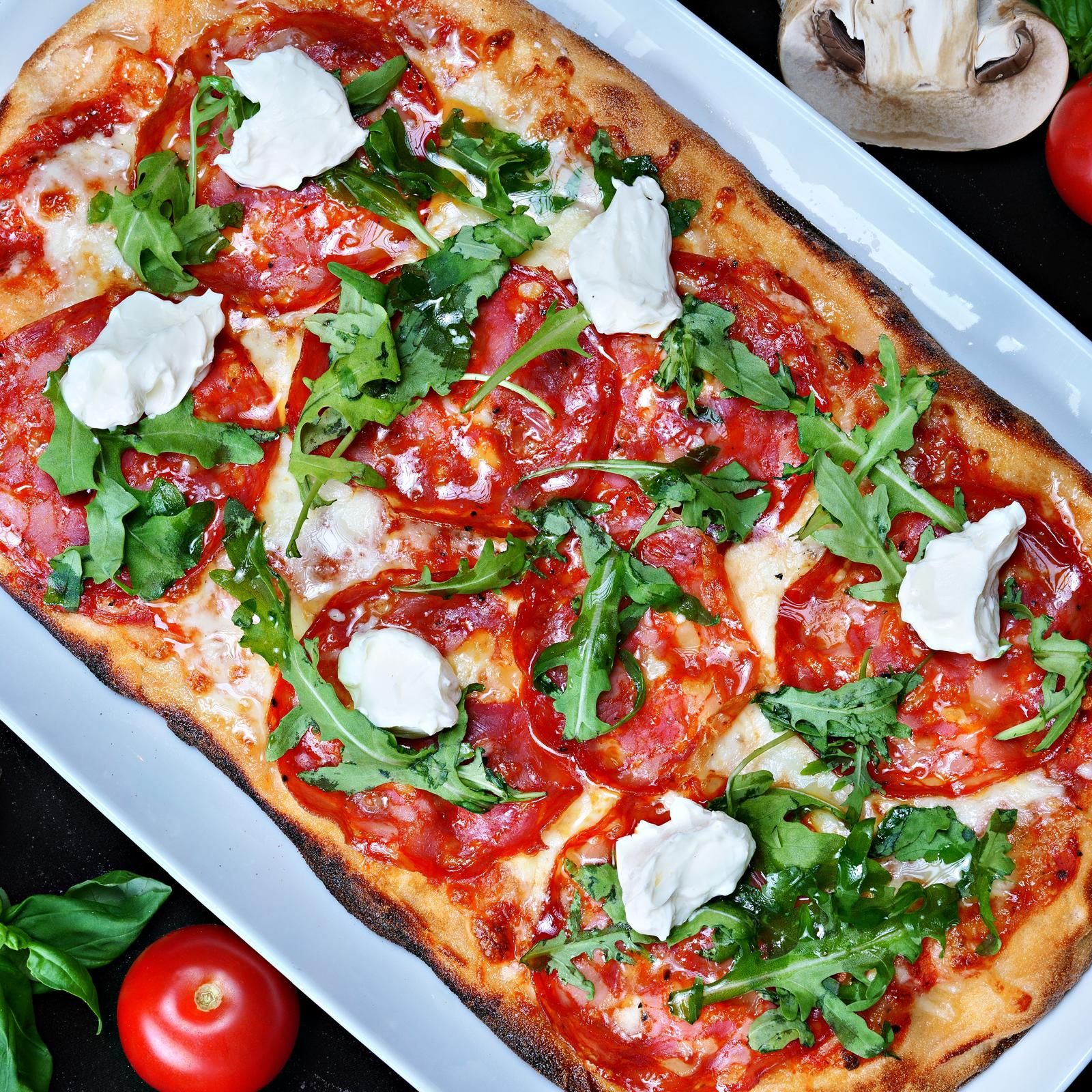 Pizza delicioasă cu felii de salam, mozzarella, roșii, rucola și mozzarella cremoasă, servită pe o farfurie albă.