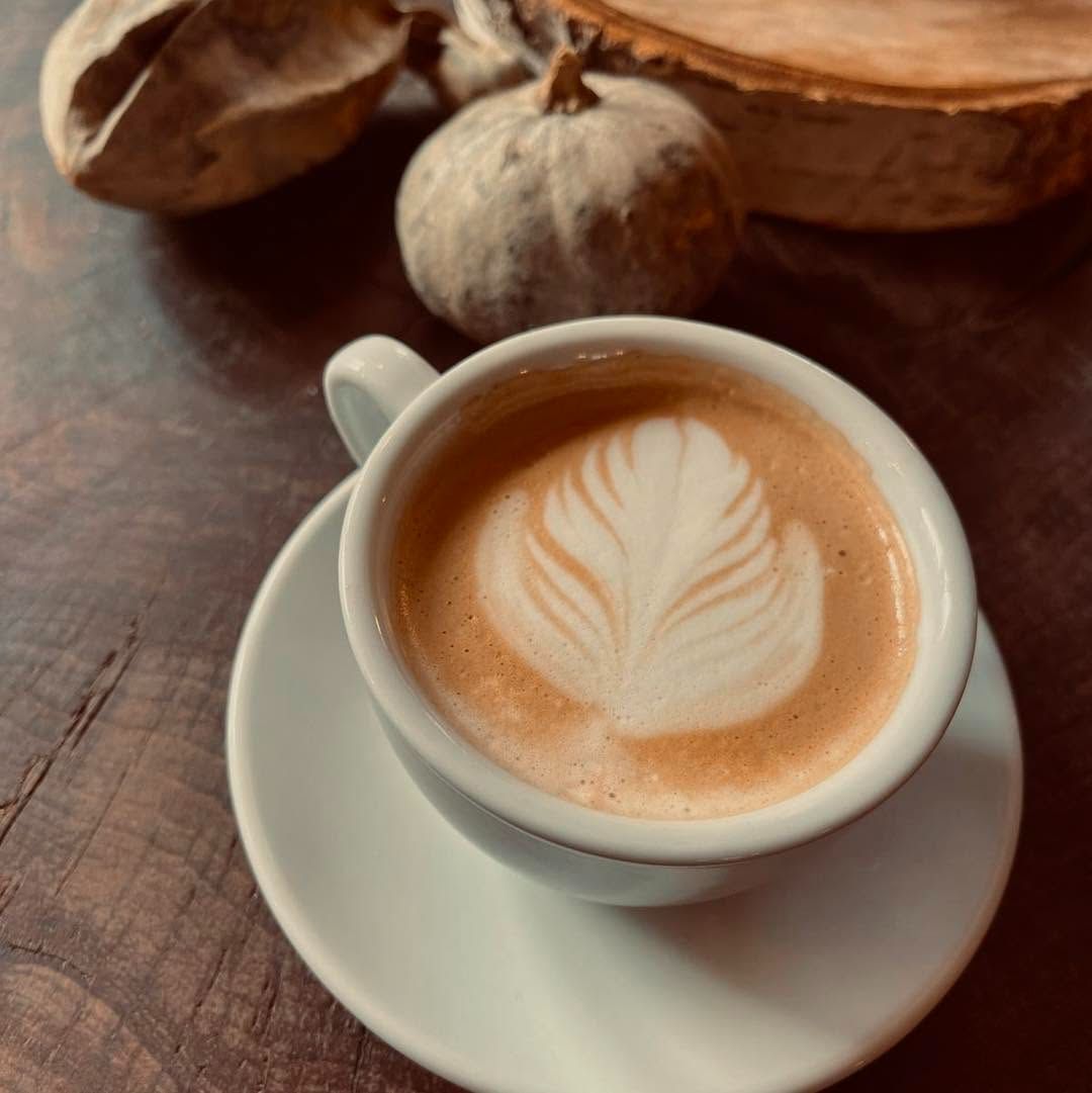 Cafetieră cu spumă de lapte în formă de frunză, alături de dovleci decorativi pe o masă din lemn.