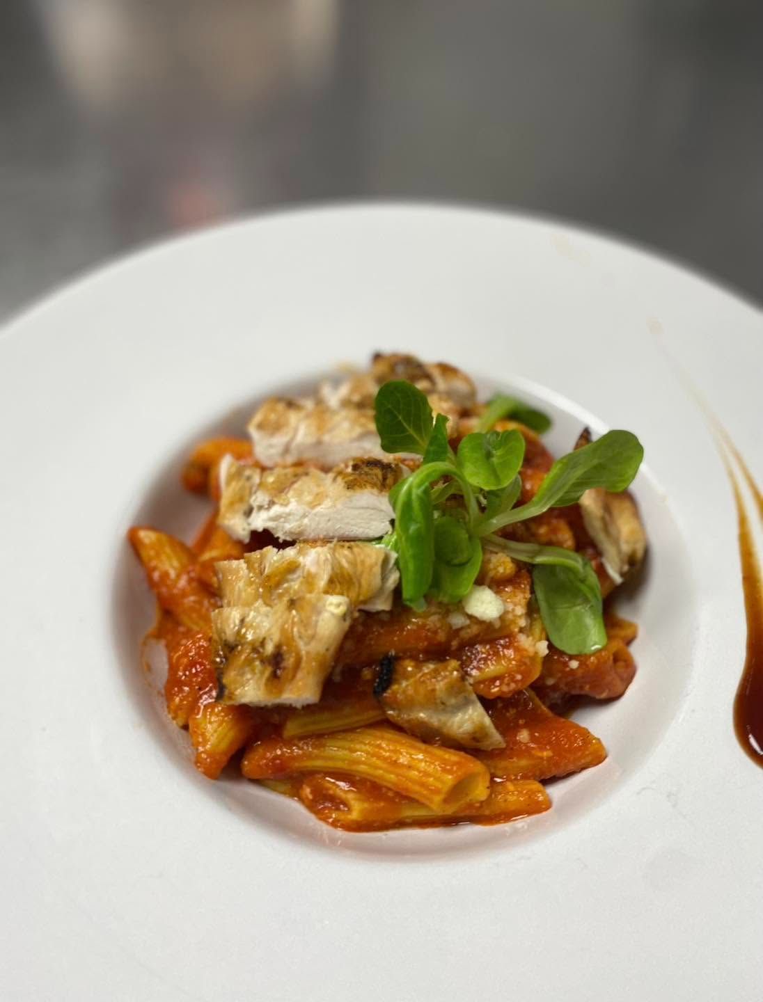 Paste penne cu sos roșu, pui la grătar și garnitură de frunze verzi, servite pe o farfurie albă.