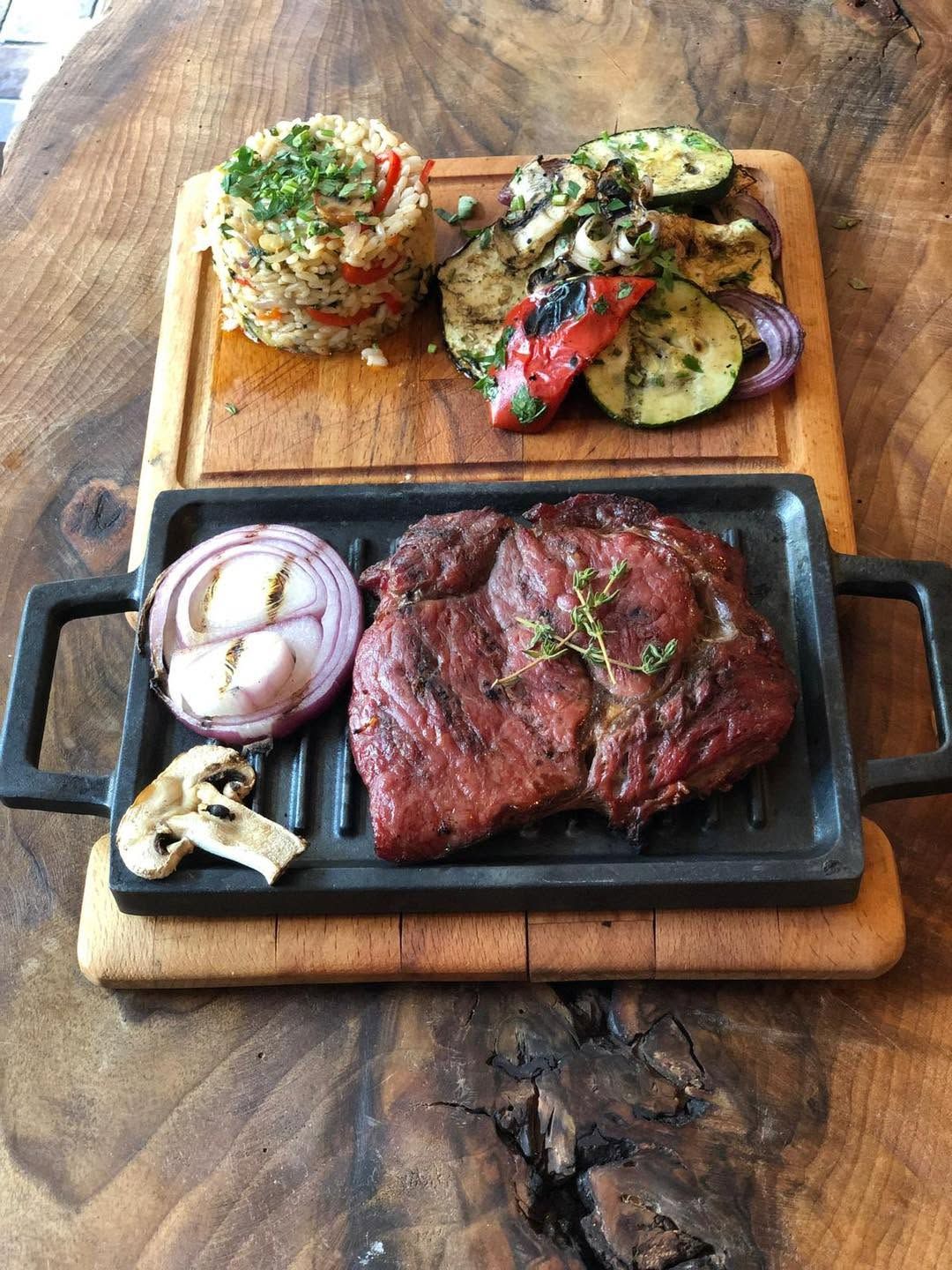 Carne de vită friptă pe un grătar, cu legume și orez pe un tocător din lemn.
