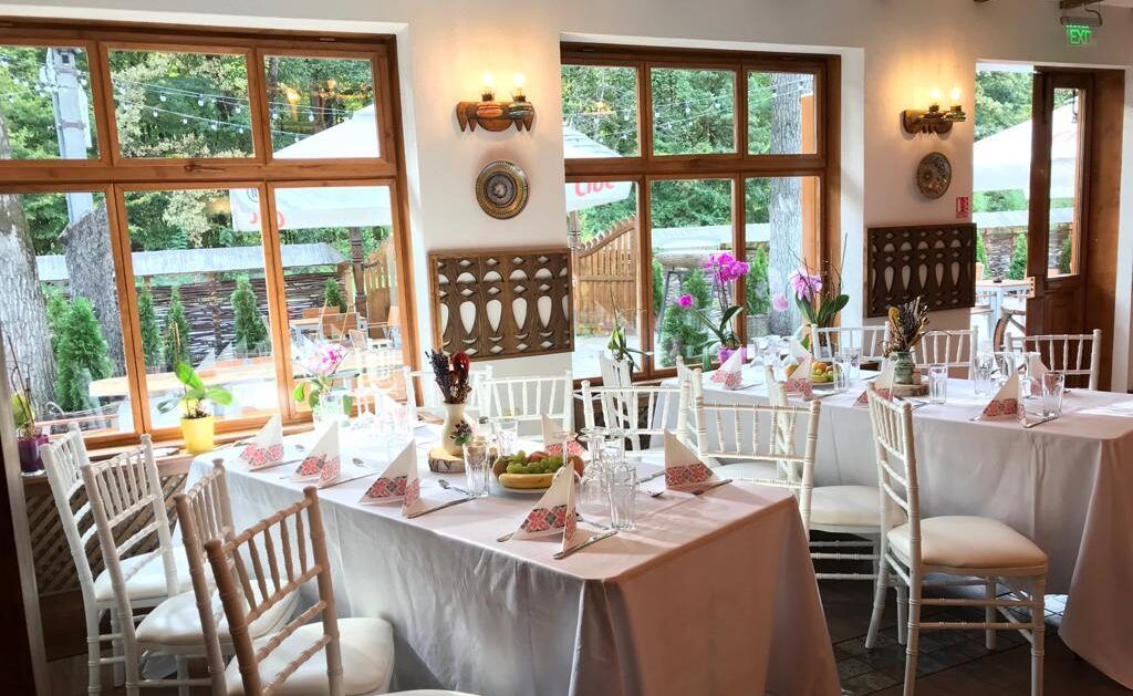 Interiorul unui restaurant elegant, cu mese acoperite cu fețe albe și flori, având feronerie din lemn și lumină naturală.
