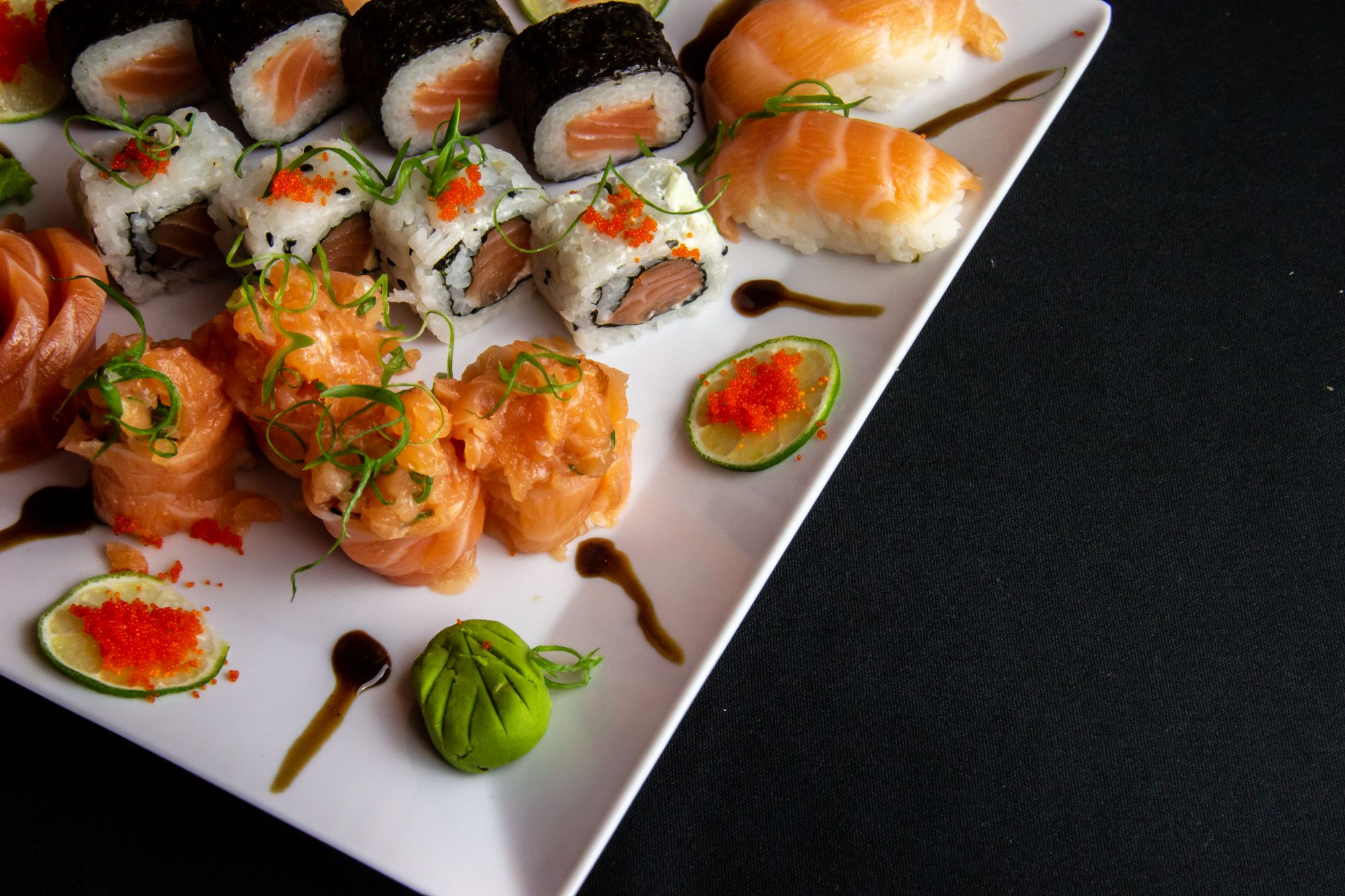Talerz sushi z różnymi rolkami, wędzonym łososiem, awokado i limonką na czarnym tle.