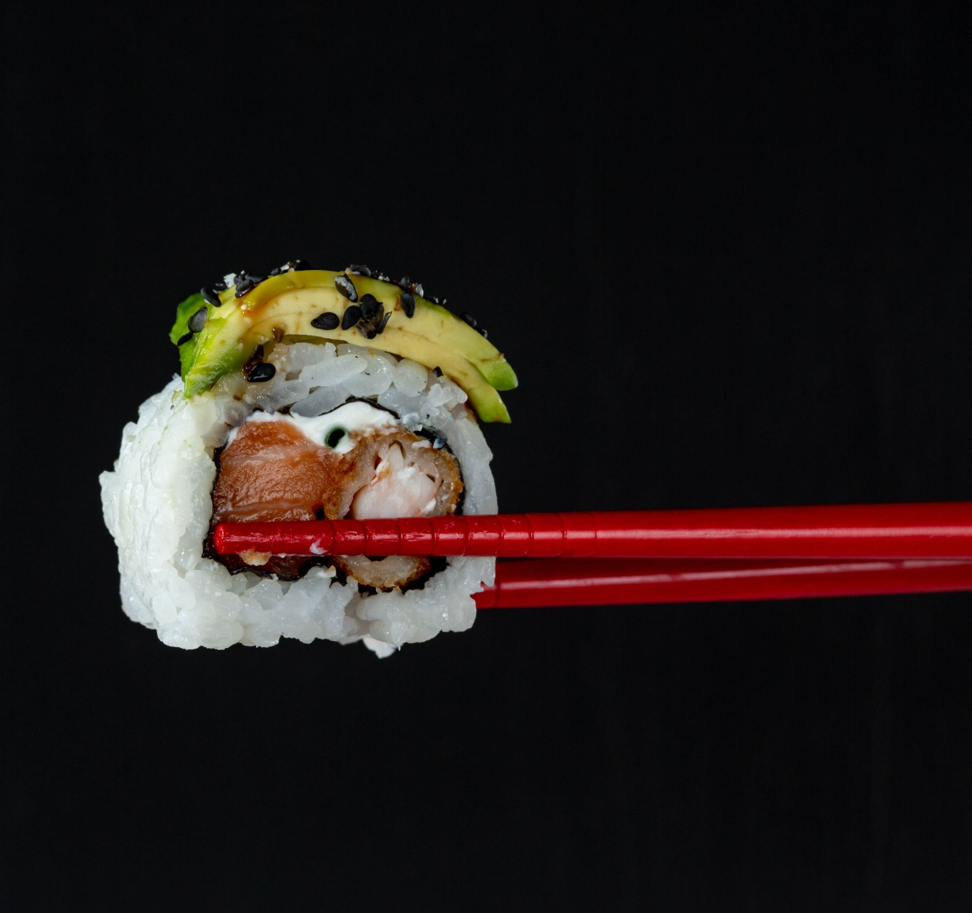 Sushi trzymane pałeczkami, z awokado, rybą i czarnym sezamem, na ciemnym tle.