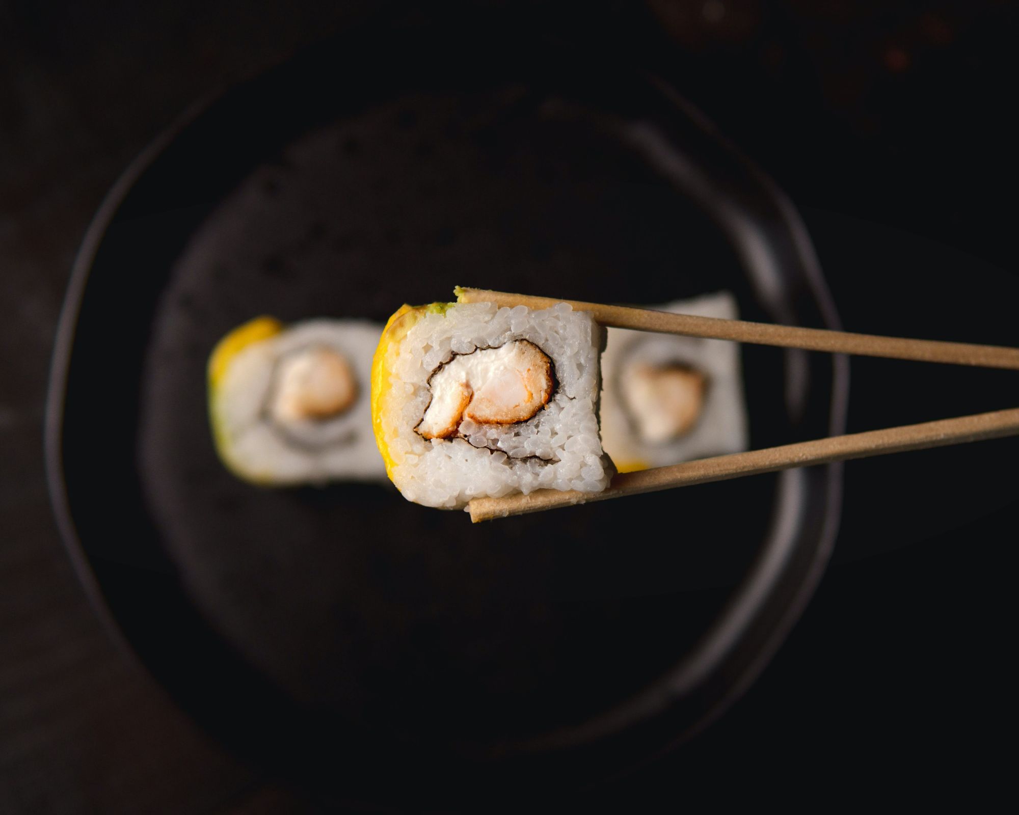 Roladki sushi trzymane w pałeczkach, na czarnym talerzu, z widoczną krewetką w środku.