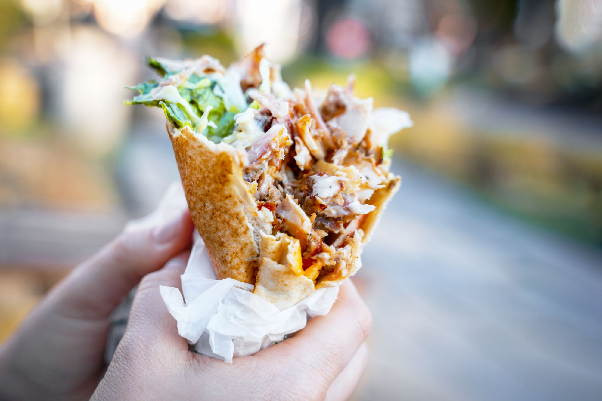 Osoba trzymająca kebab w bułce z zielonymi liśćmi na tle miejskiego krajobrazu.