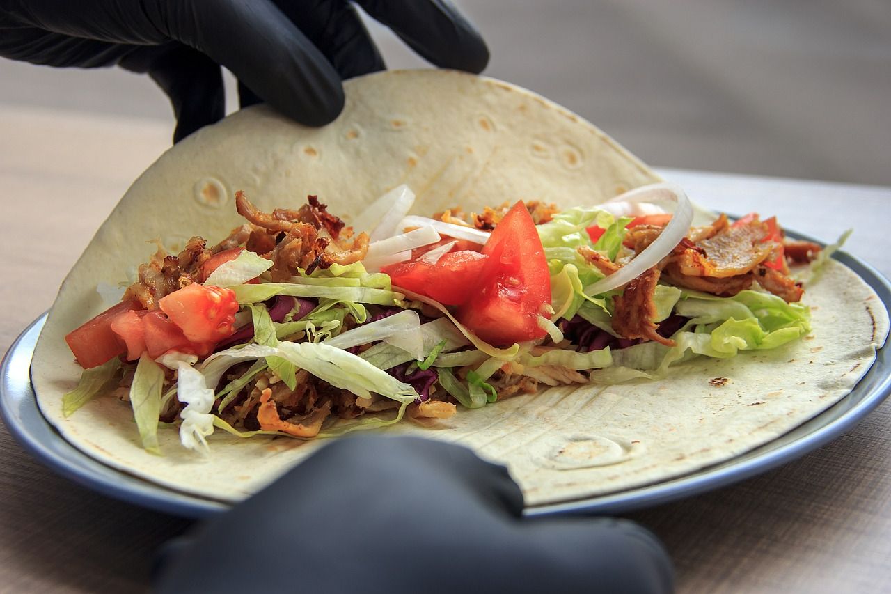 Taco z kurczakiem, sałatą, pomidorem i cebulą w rękach osoby w czarnych rękawiczkach.
