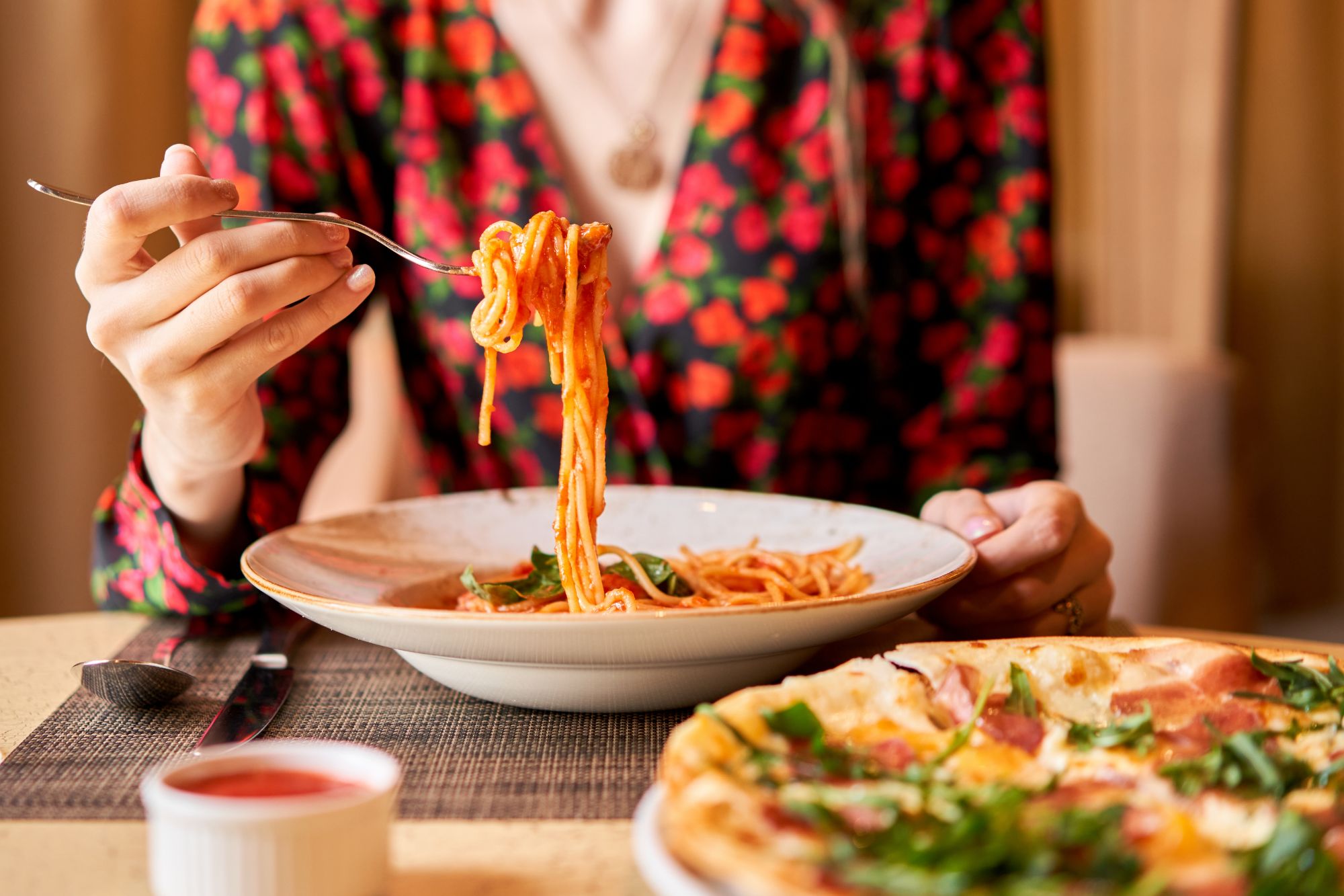 Osoba w kwiecistej bluzce trzyma widelec z makaronem, obok talerz spaghetti i pizza.