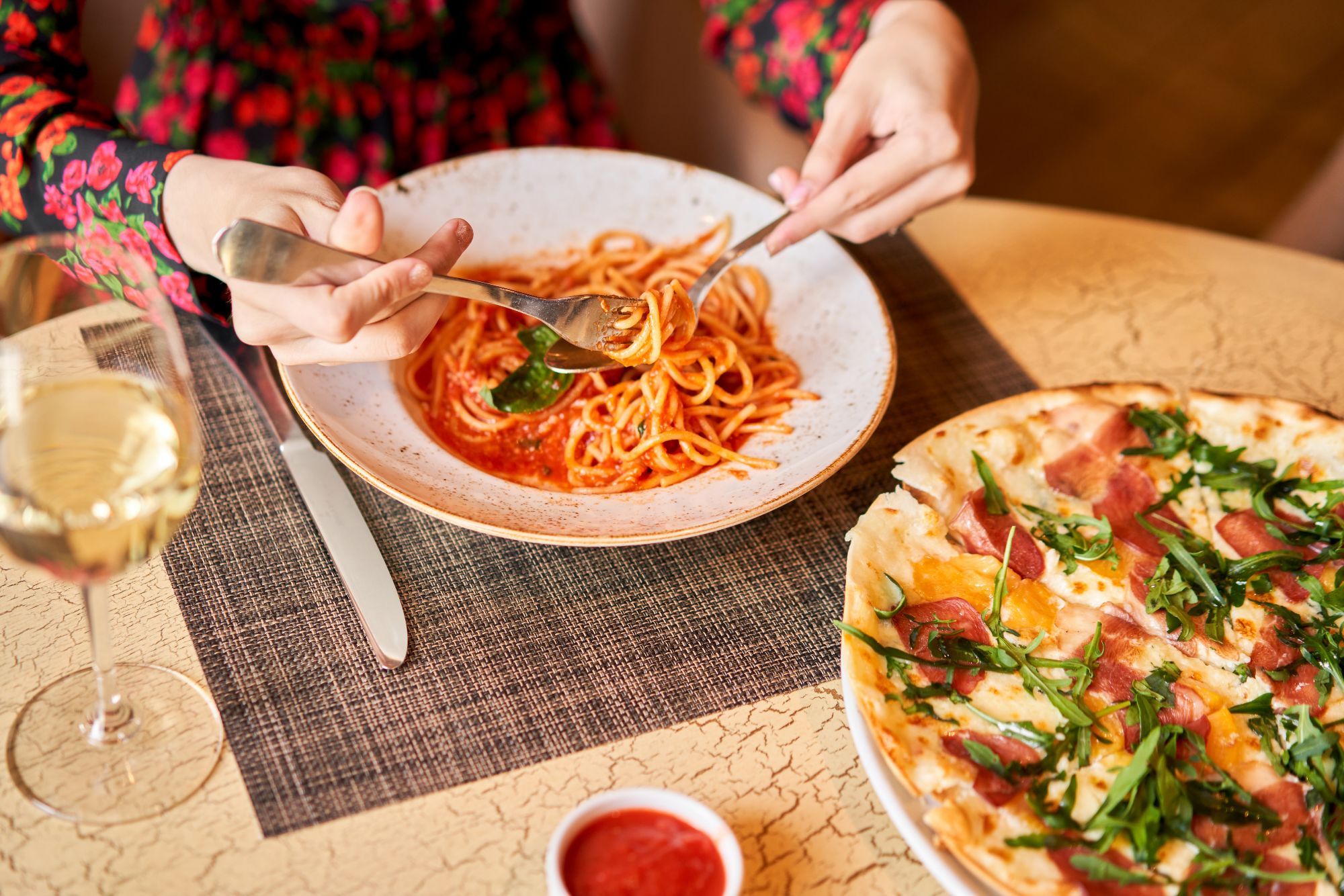Kobieta je spaghetti w sosie pomidorowym, obok pizza z dodatkami i kieliszek białego wina na stole.