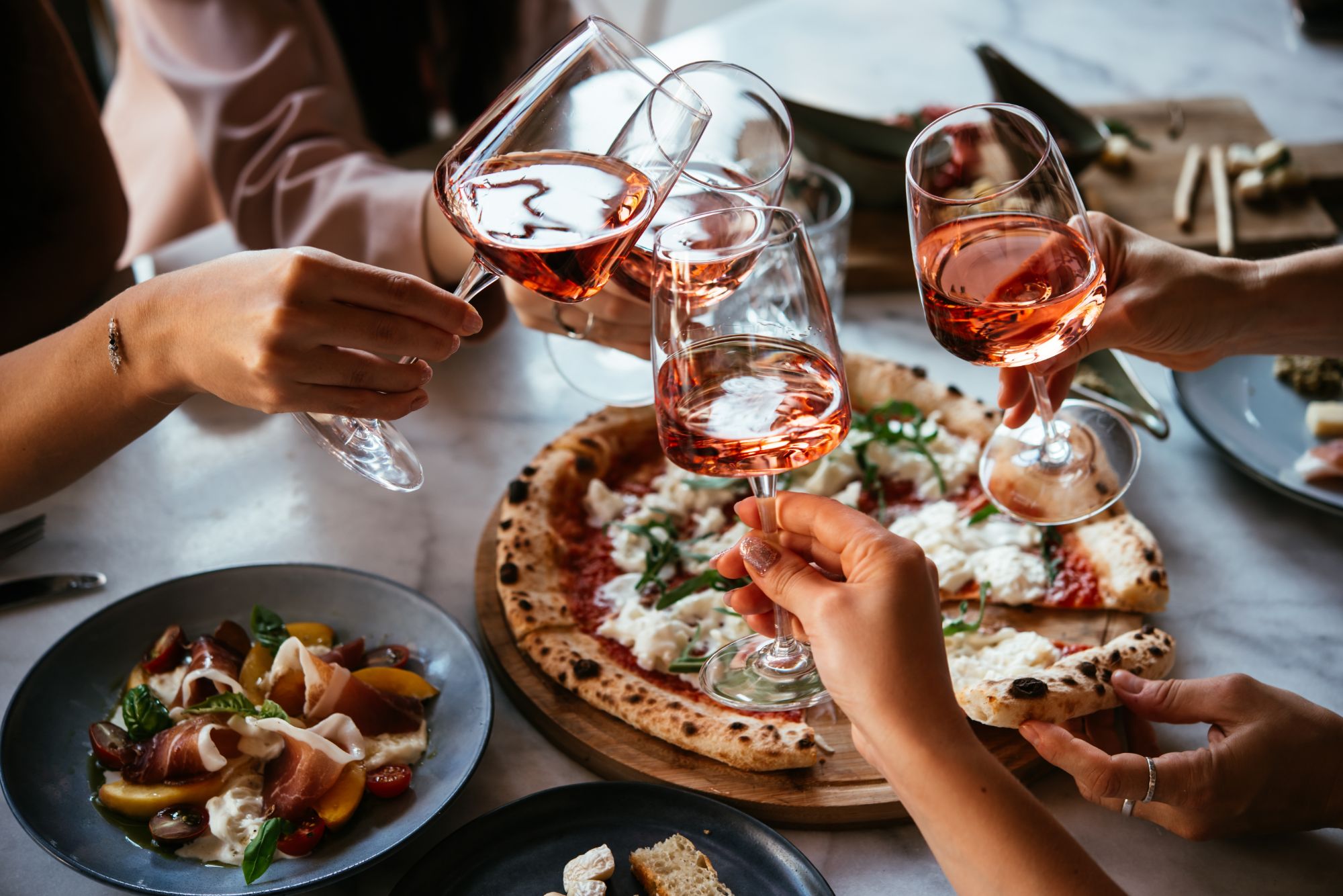 Zbliżenie na ręce unoszące kieliszki różowego wina nad pizzą i sałatką na stole.