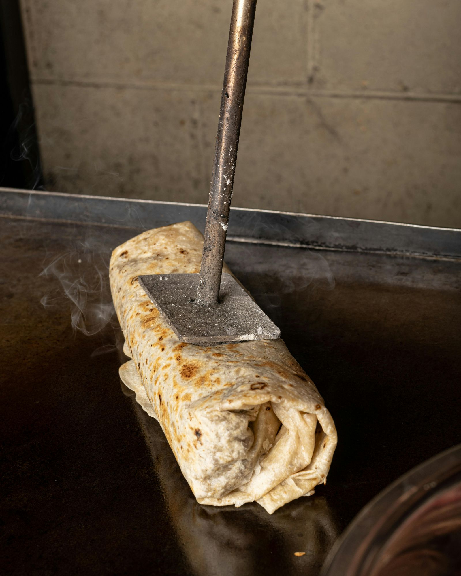Smażona tortilla na grillu z dymiącym nadzieniem, przyciskana metalowym narzędziem.