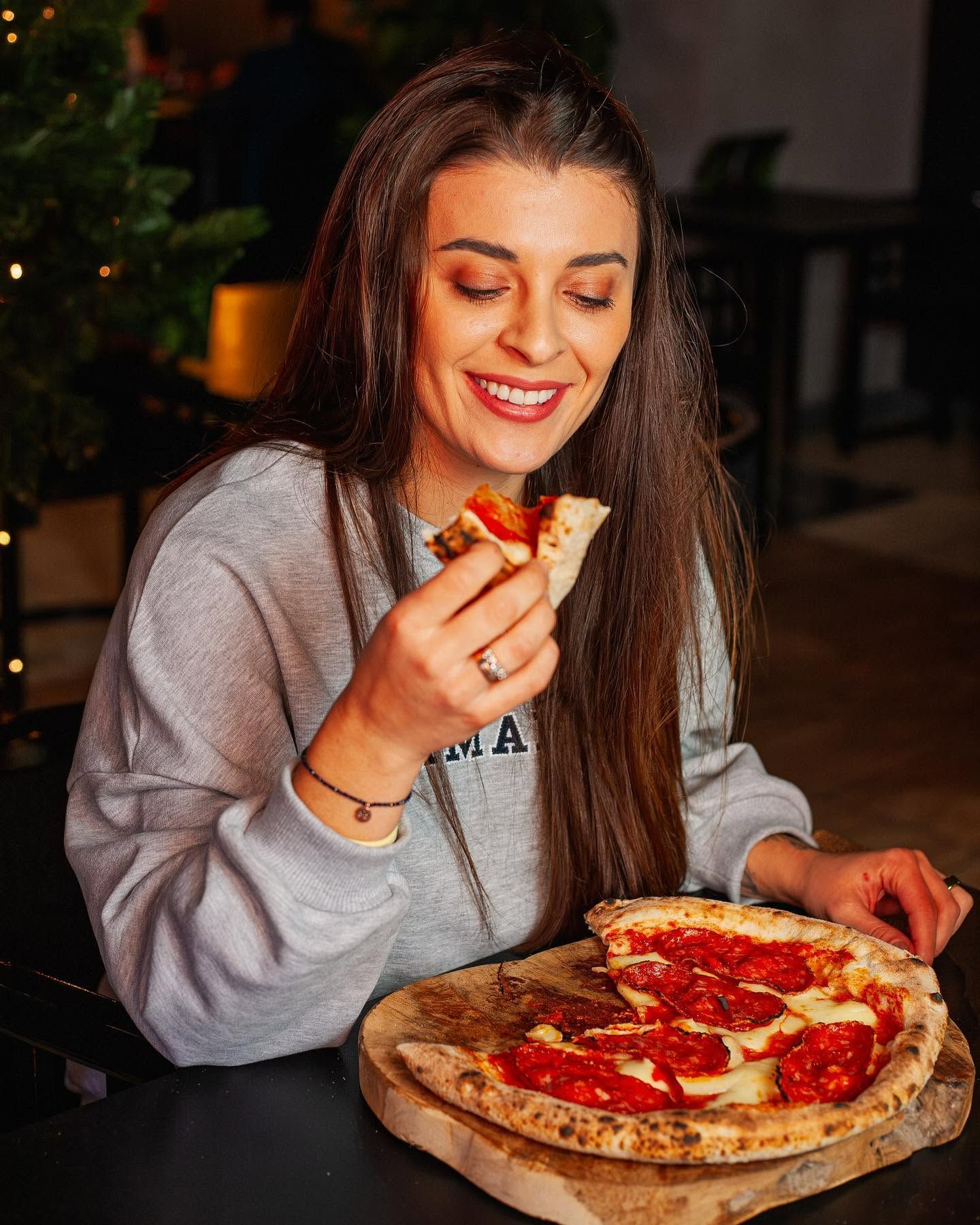 Kobieta z długimi włosami uśmiecha się, jedząc pizzę z pepperoni w przytulnej restauracji.