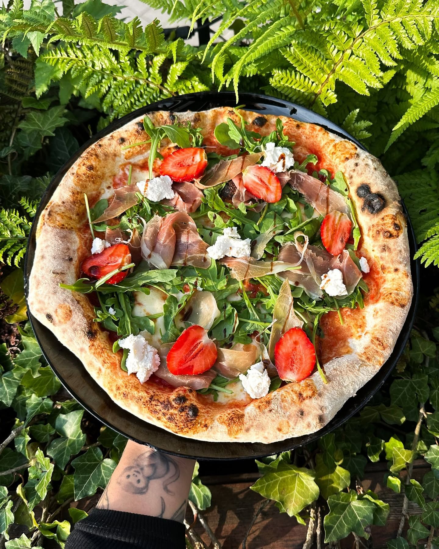 Pizza z rukolą, pomidorami, szynką, serem kozimi na czarnym talerzu, otoczona zielenią.