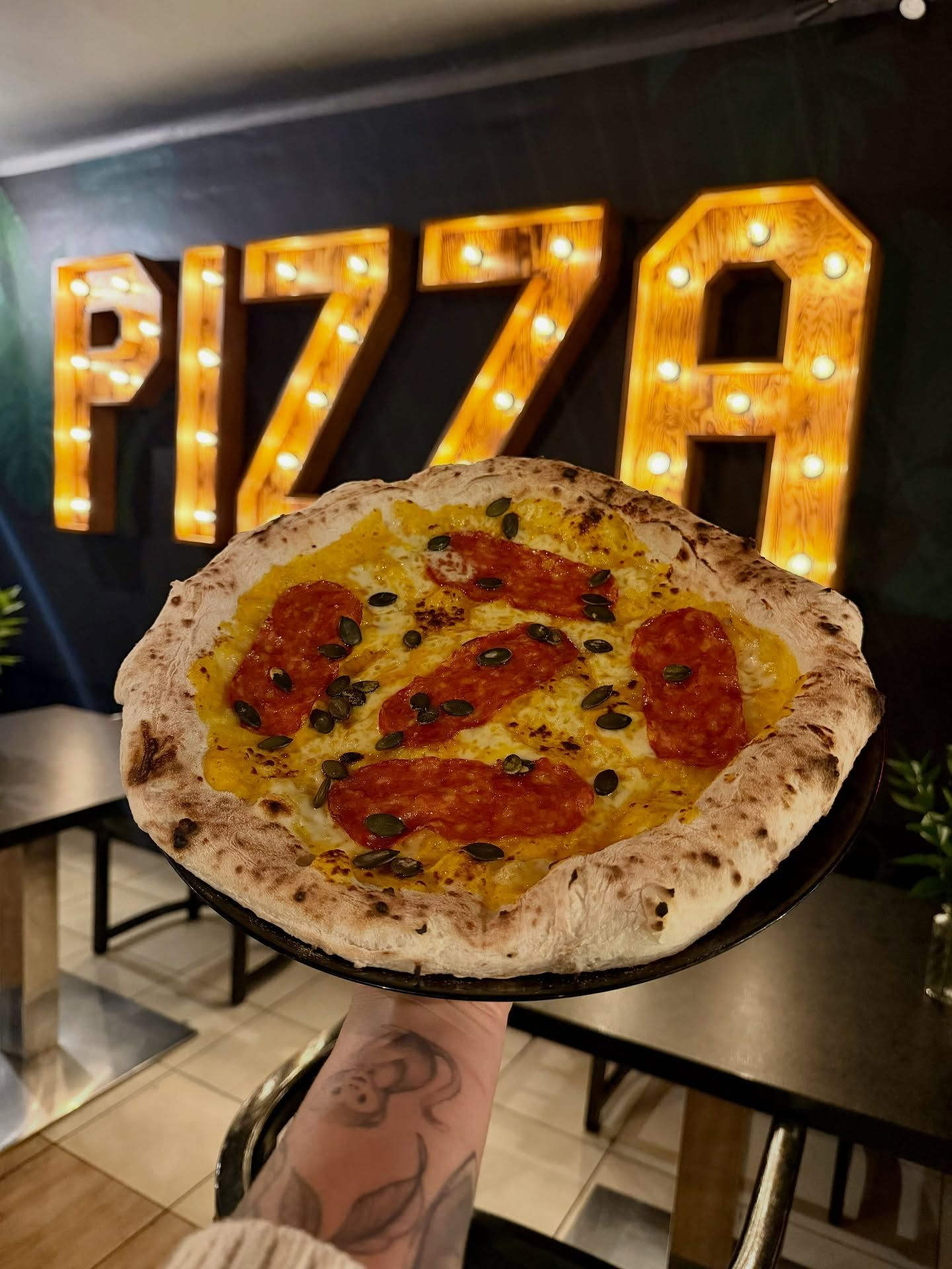 Pizza z pepperoni trzymana w dłoni, w tle neonowy napis „PIZZA”.