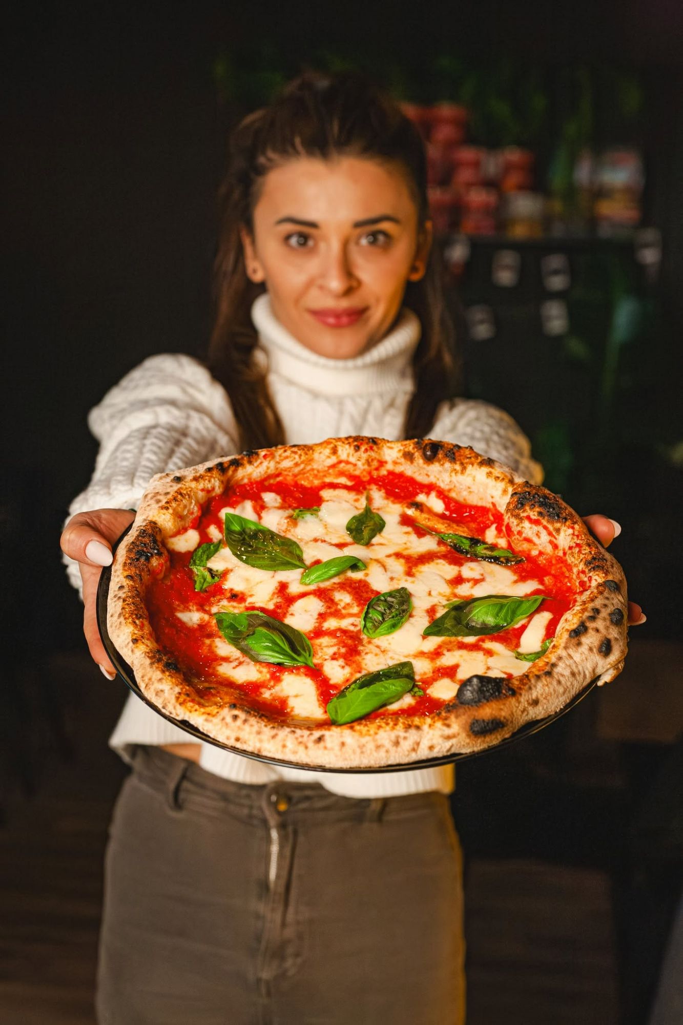 Kobieta trzyma na talerzu pizzę margherita z bazylią, z uśmiechem w przytulnej restauracji.