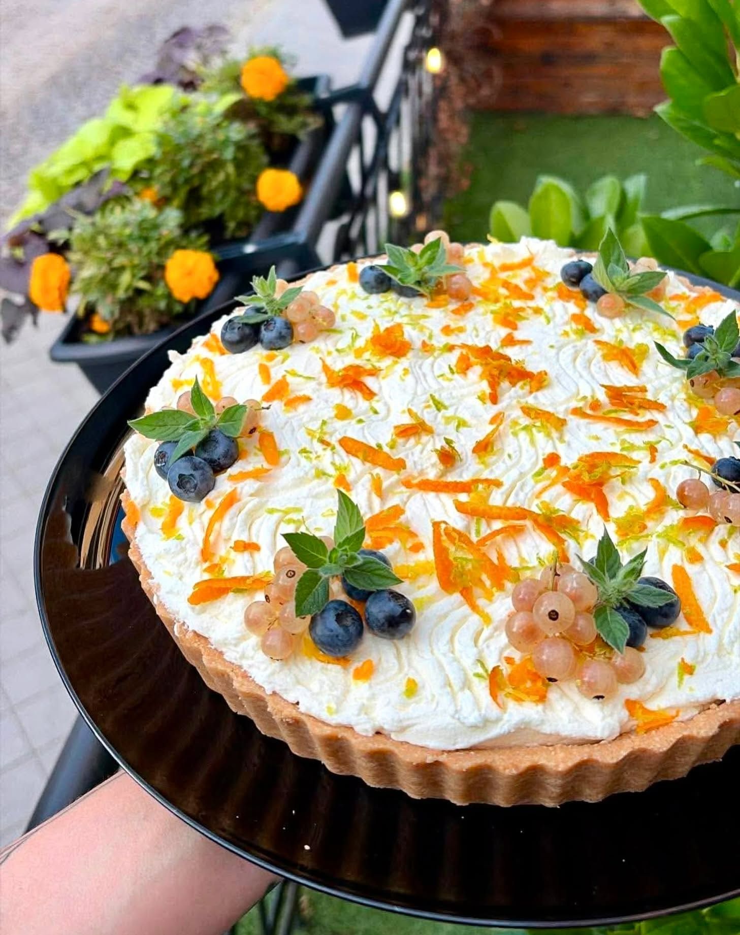 Tarta z kremem, dekorowana jagodami, porzeczkami i miętą, na czarnej paterze. W tle kwiaty na balkonie.