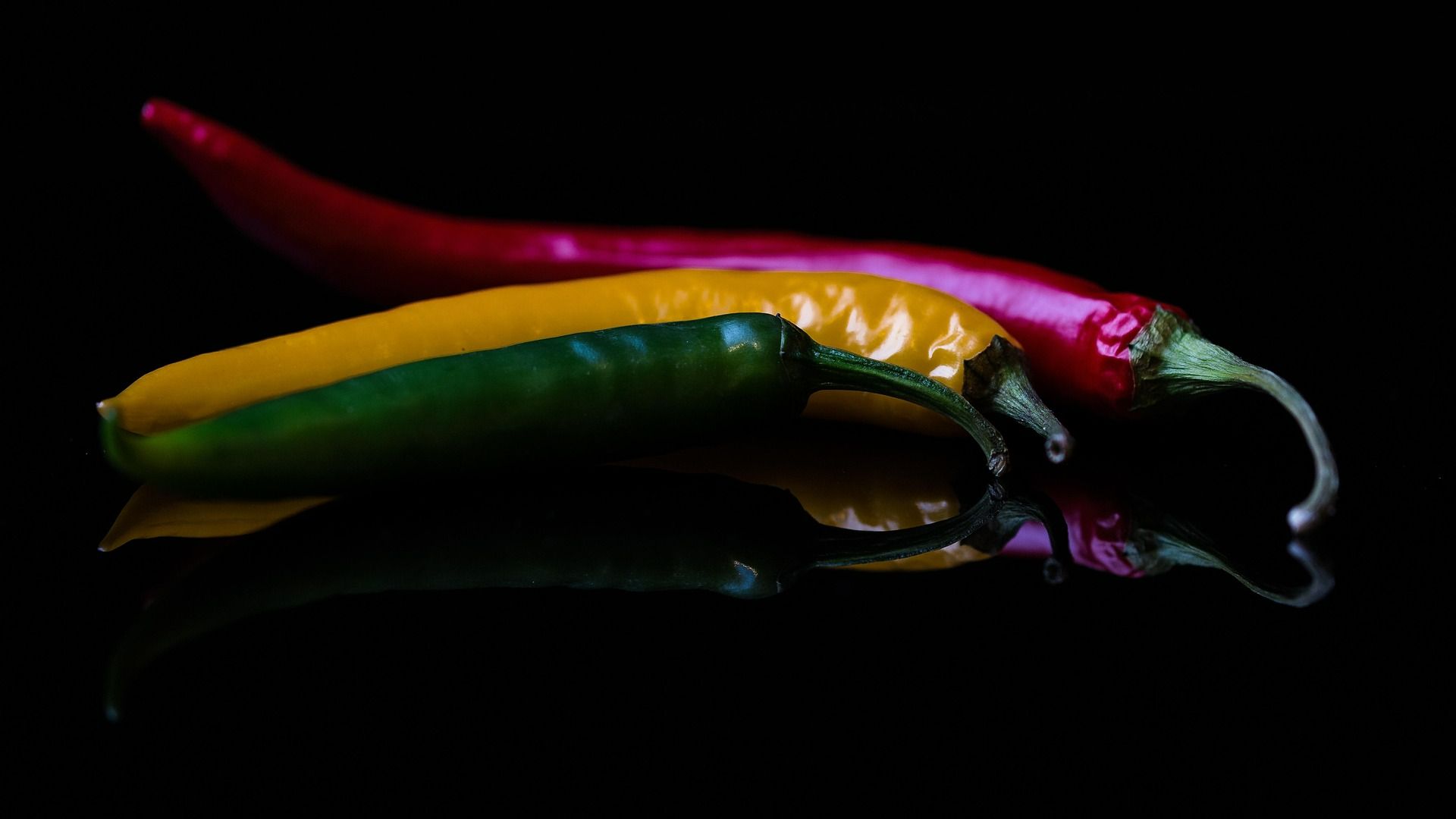 Trzy różnokolorowe papryki chili: czerwona, żółta i zielona, na czarnym tle z odbiciem.
