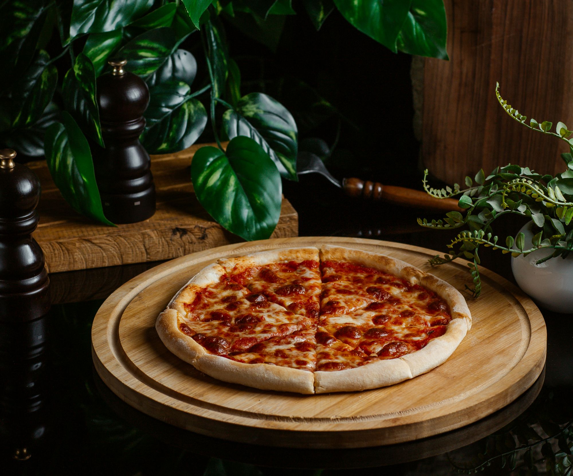 Pizza pepperoni na drewnianym talerzu, w tle zielone rośliny i przyprawniki.