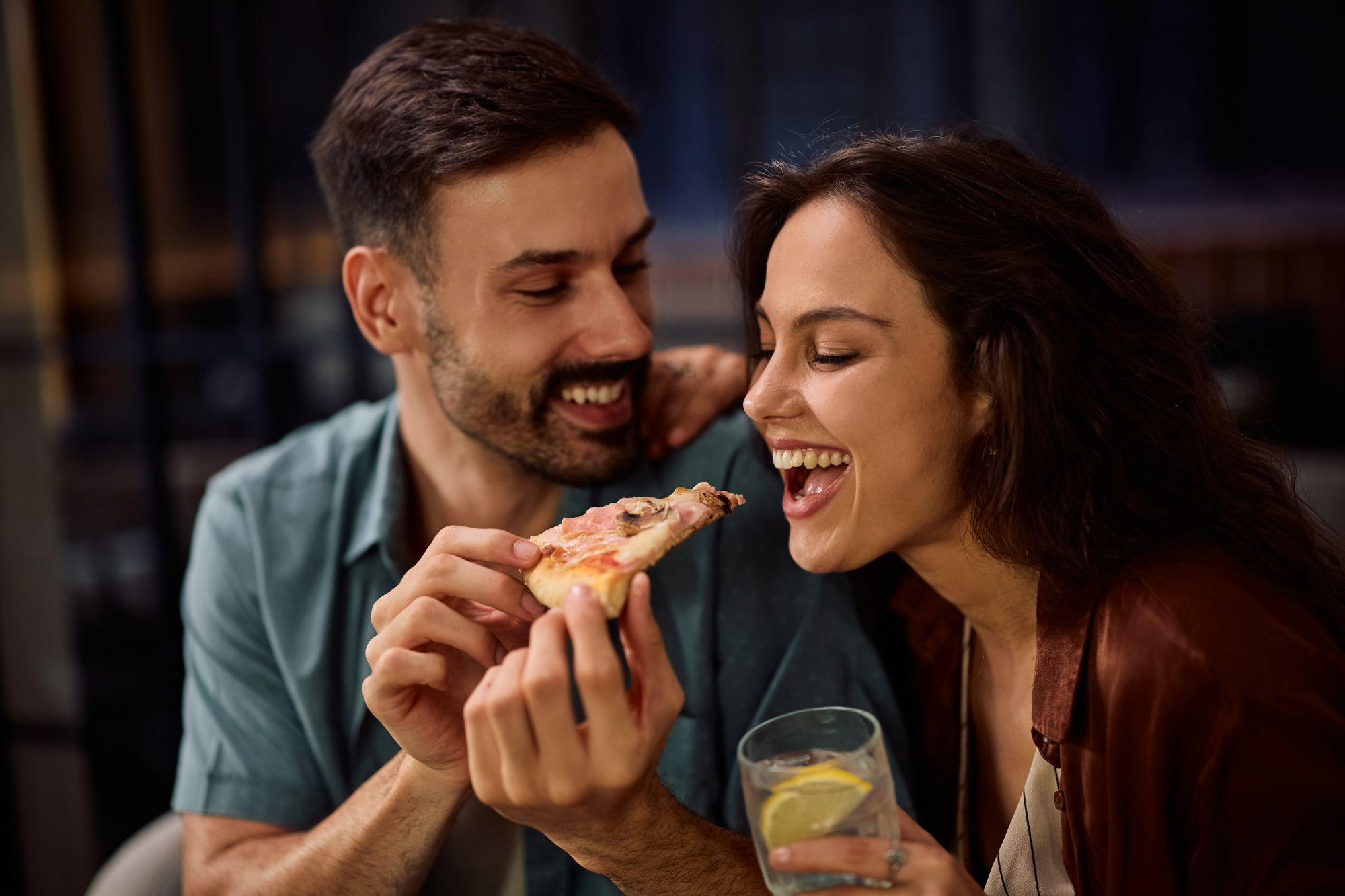 Mężczyzna i kobieta uśmiechają się, dzieląc się kawałkiem pizzy przy romantycznej kolacji.