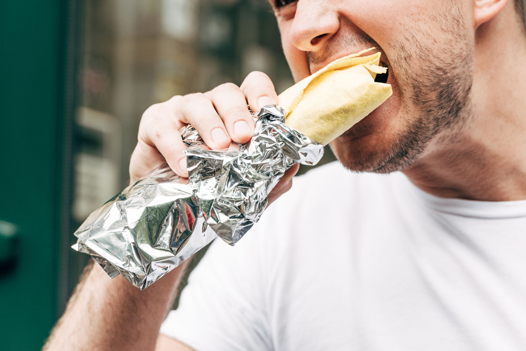 Mężczyzna w białej koszulce gryzie burrito owinięte w folię aluminiową.
