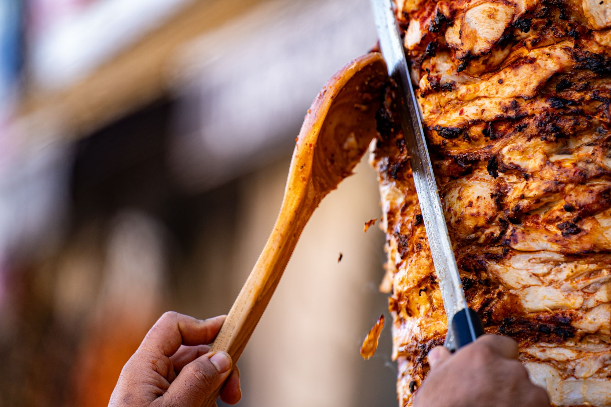 Osoba kroi soczysty kebab na rożnie za pomocą drewnianej łyżki i dużego noża.