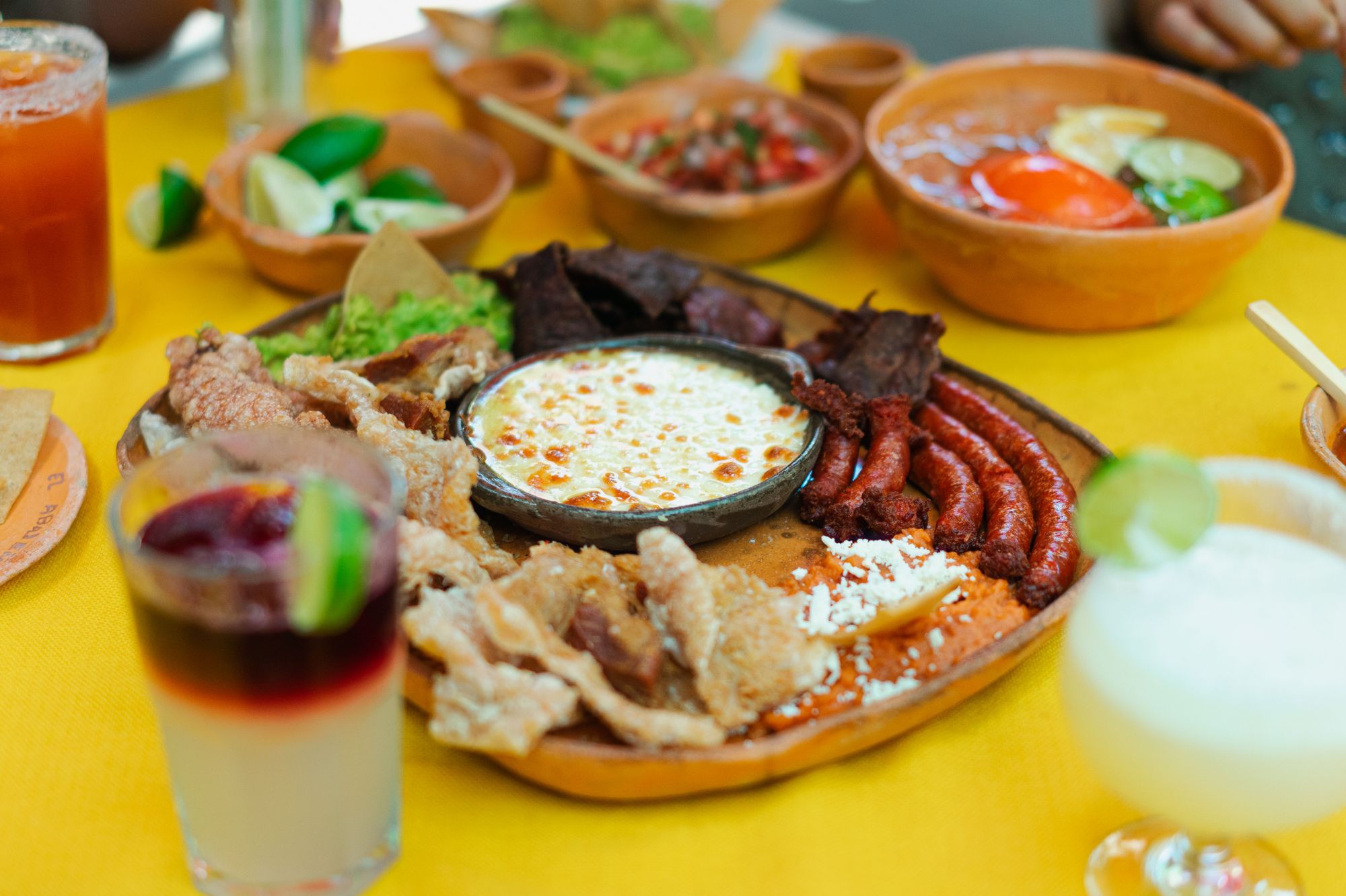 Taca z potrawami: mięso, ser, salsa, tortilla, przekąski i napoje na żółtym stole.