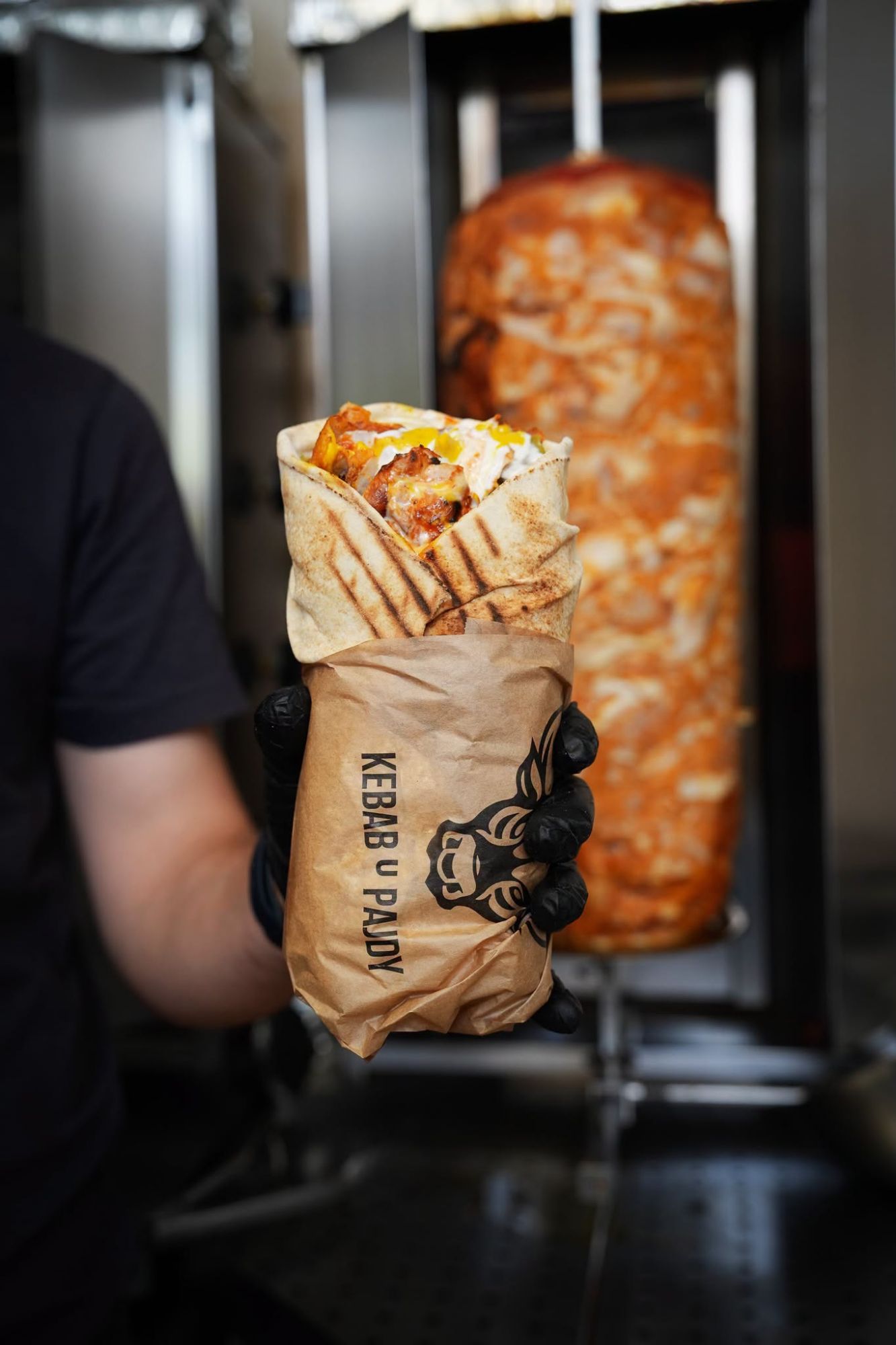 Ręka trzyma kebab zawinięty w papier, w tle obrócony szaszłyk z mięsem.