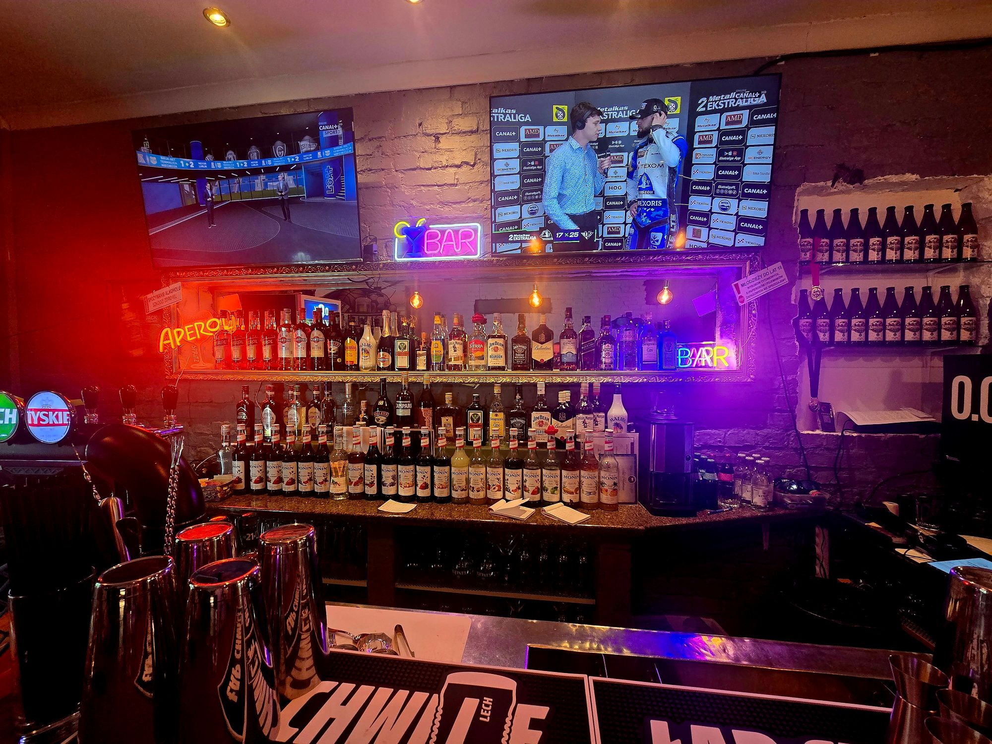 Widok na bar z butelkami alkoholu, neonowymi znakami i telewizorami w tle.