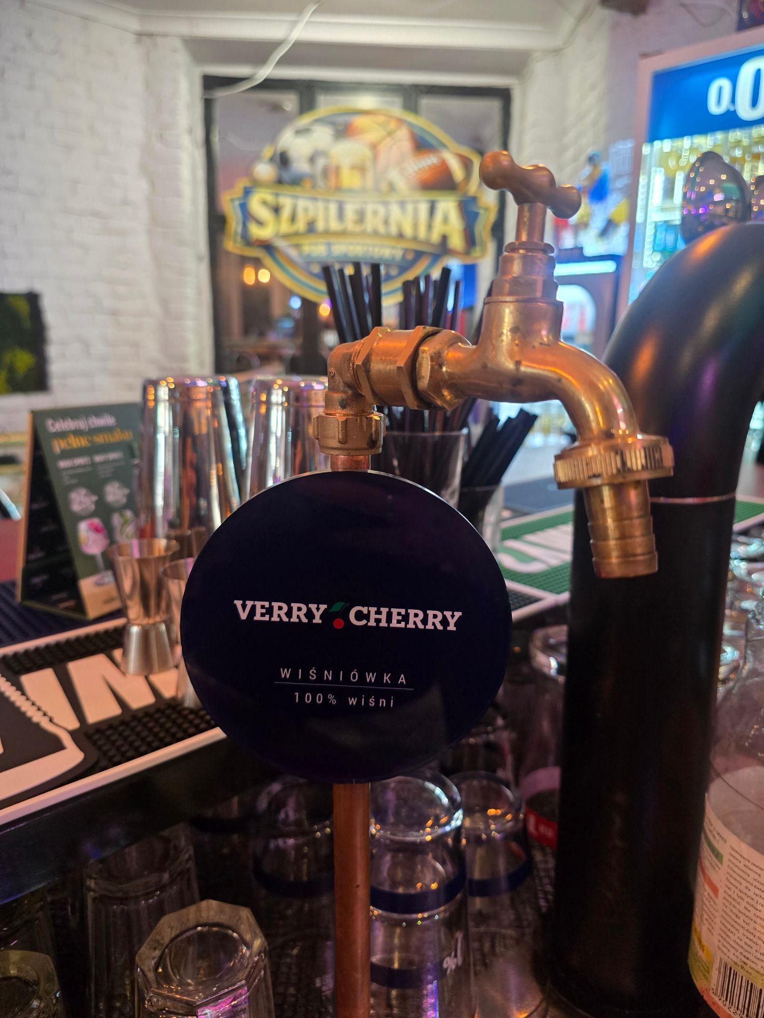 Kran z napojem VERRY CHERRY, wisniówką 100% wiśni, w barze z muralem Szpilernia w tle.