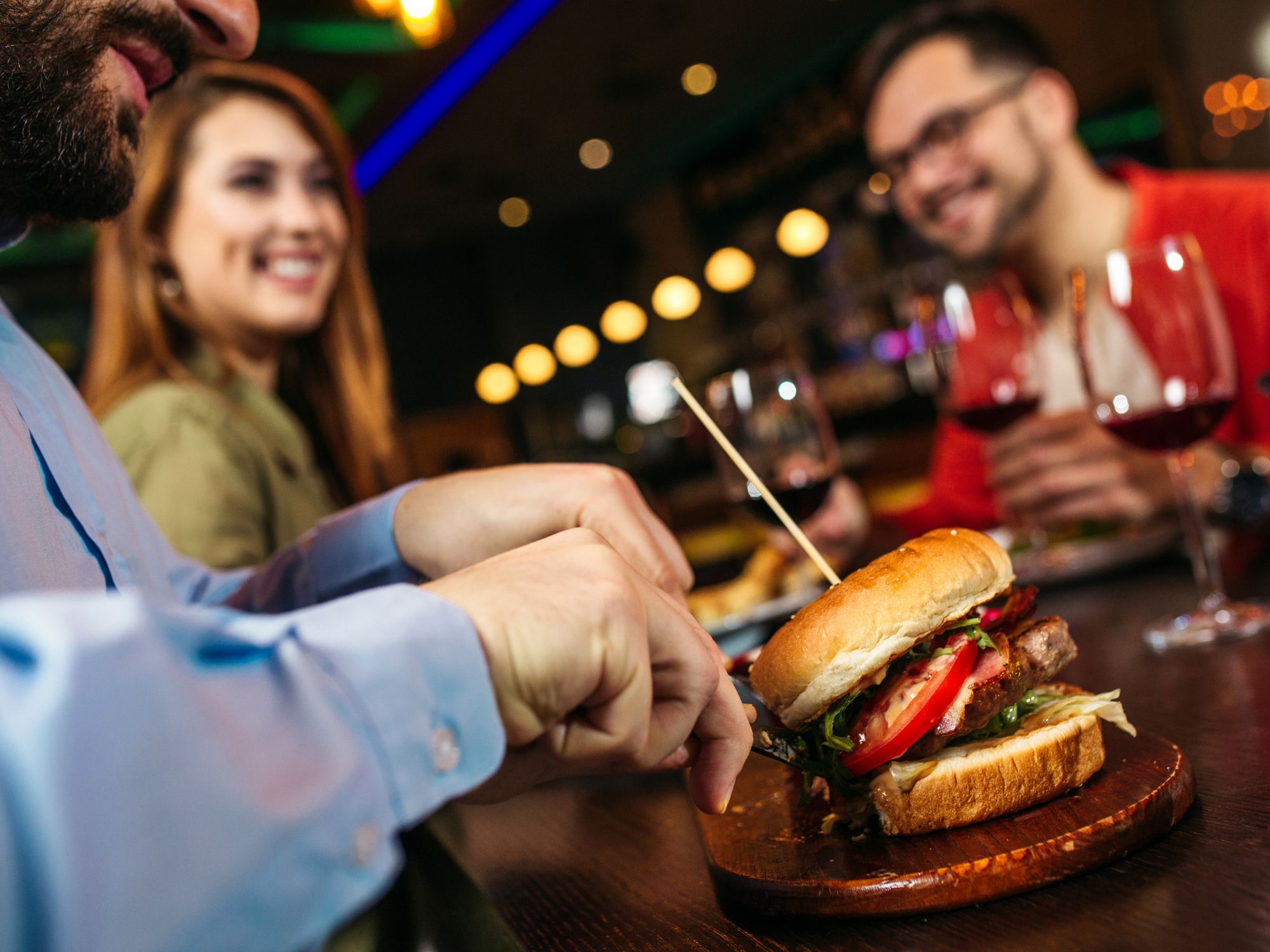 Osoba kroi hamburgera w restauracji, w tle uśmiechnięci znajomi z kieliszkami wina.