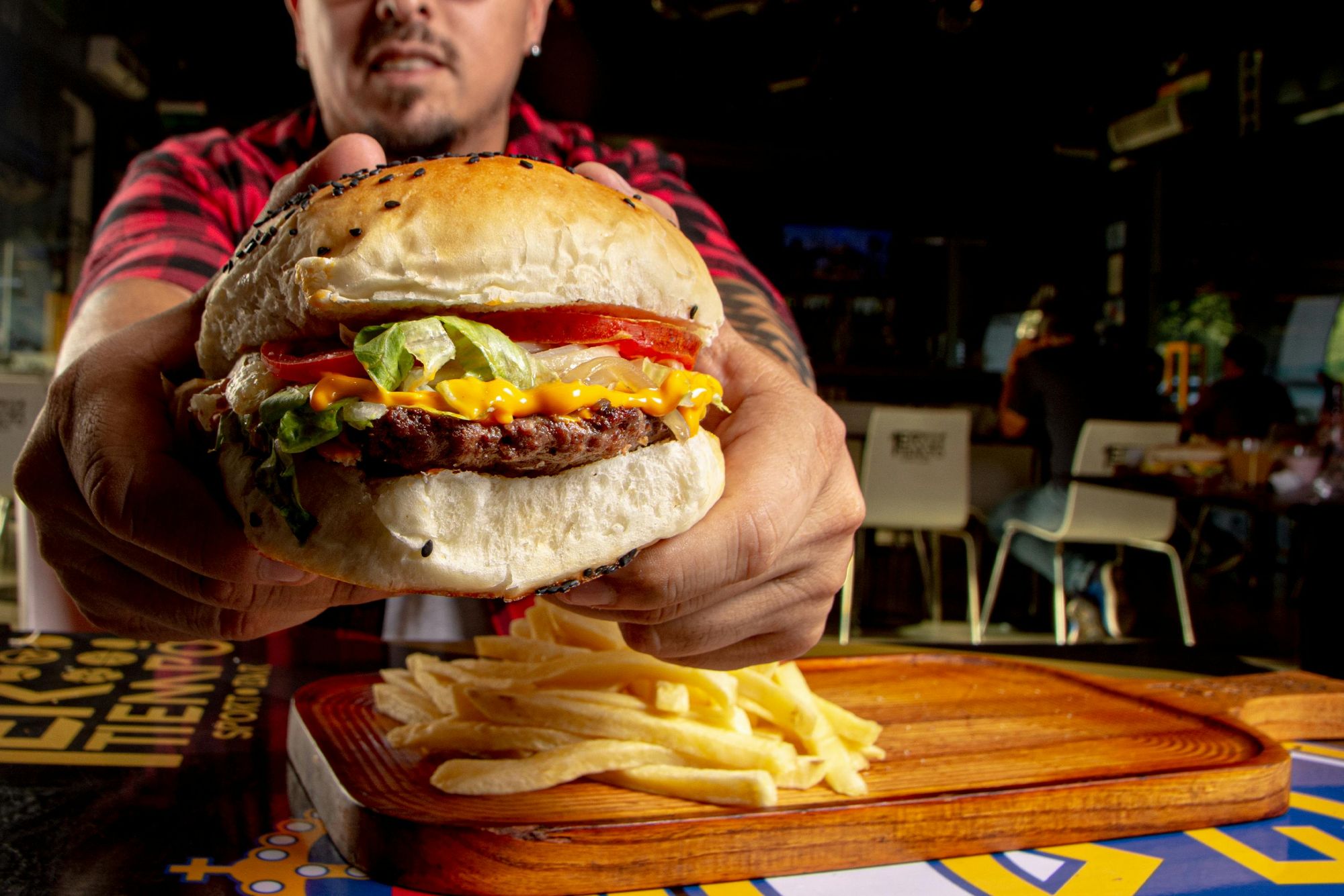 Mężczyzna trzyma dużego burgera z sałatą, pomidorem, serem i frytkami na drewnianej desce.