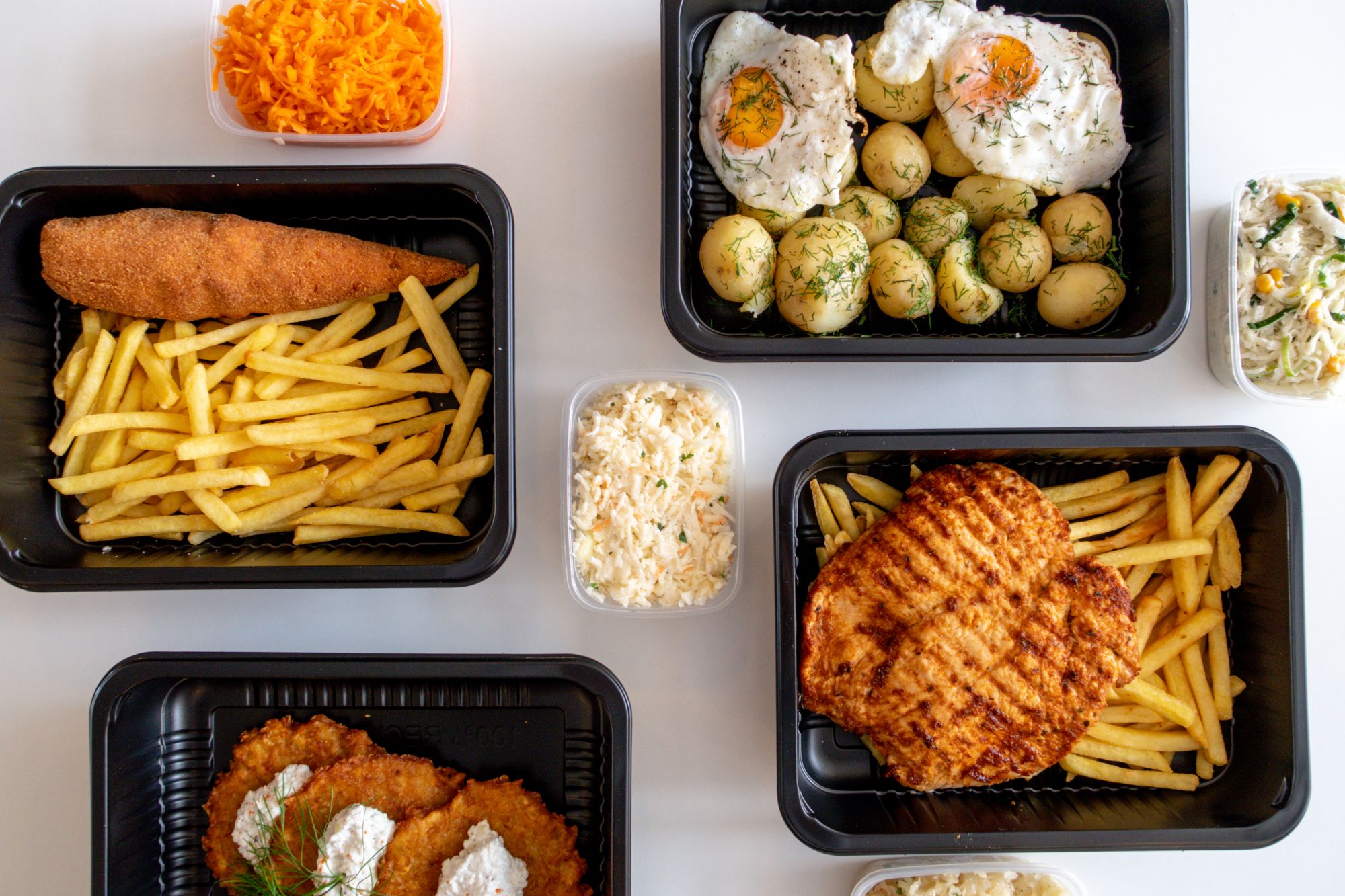 Zestaw dań: frytki, kotlet, sałatka, ziemniaki, jajka i surówki w czarnych pojemnikach.