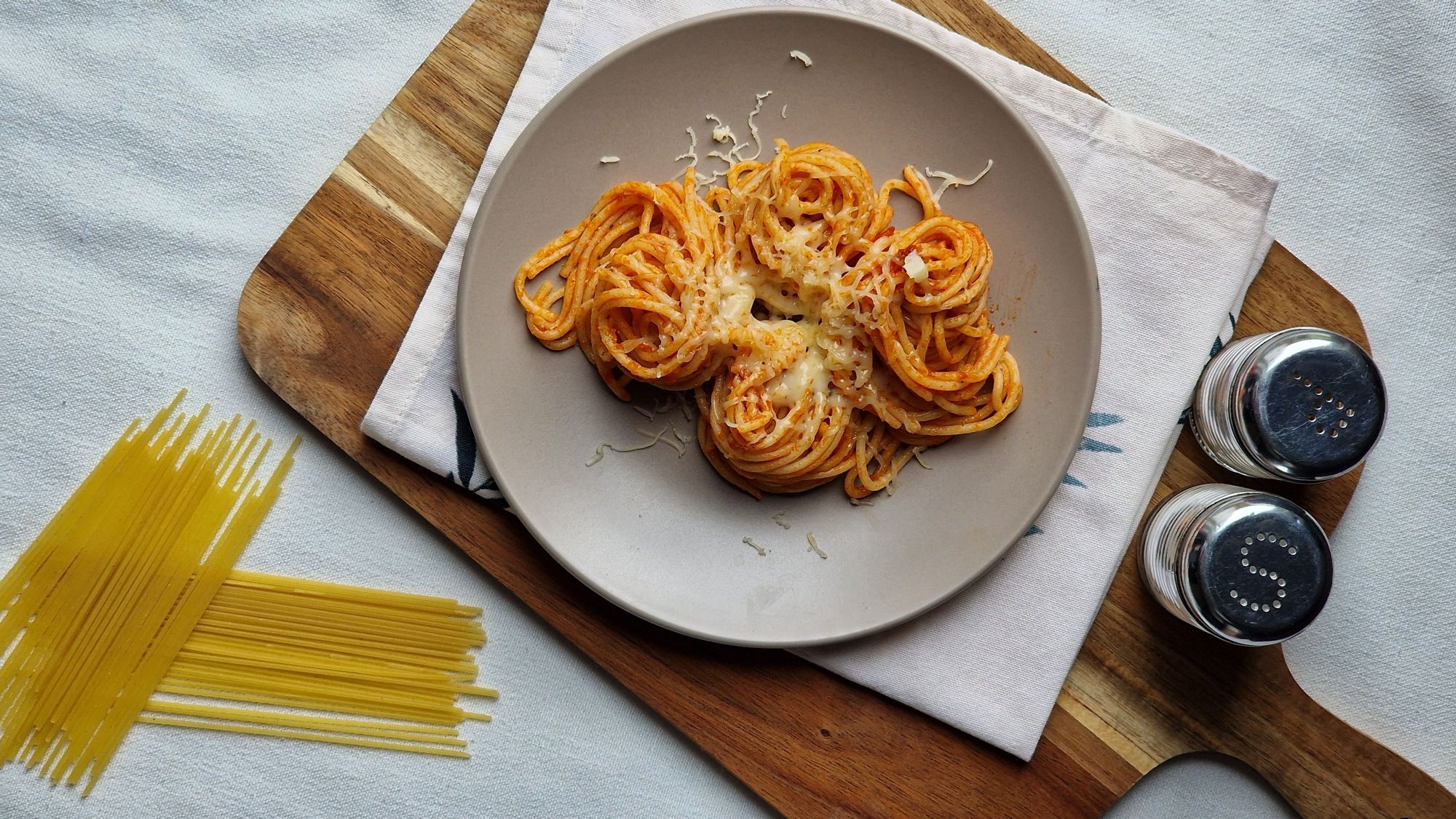 Talerz spaghetti z serem na drewnianej desce, obok sól i pieprz oraz surowy makaron.