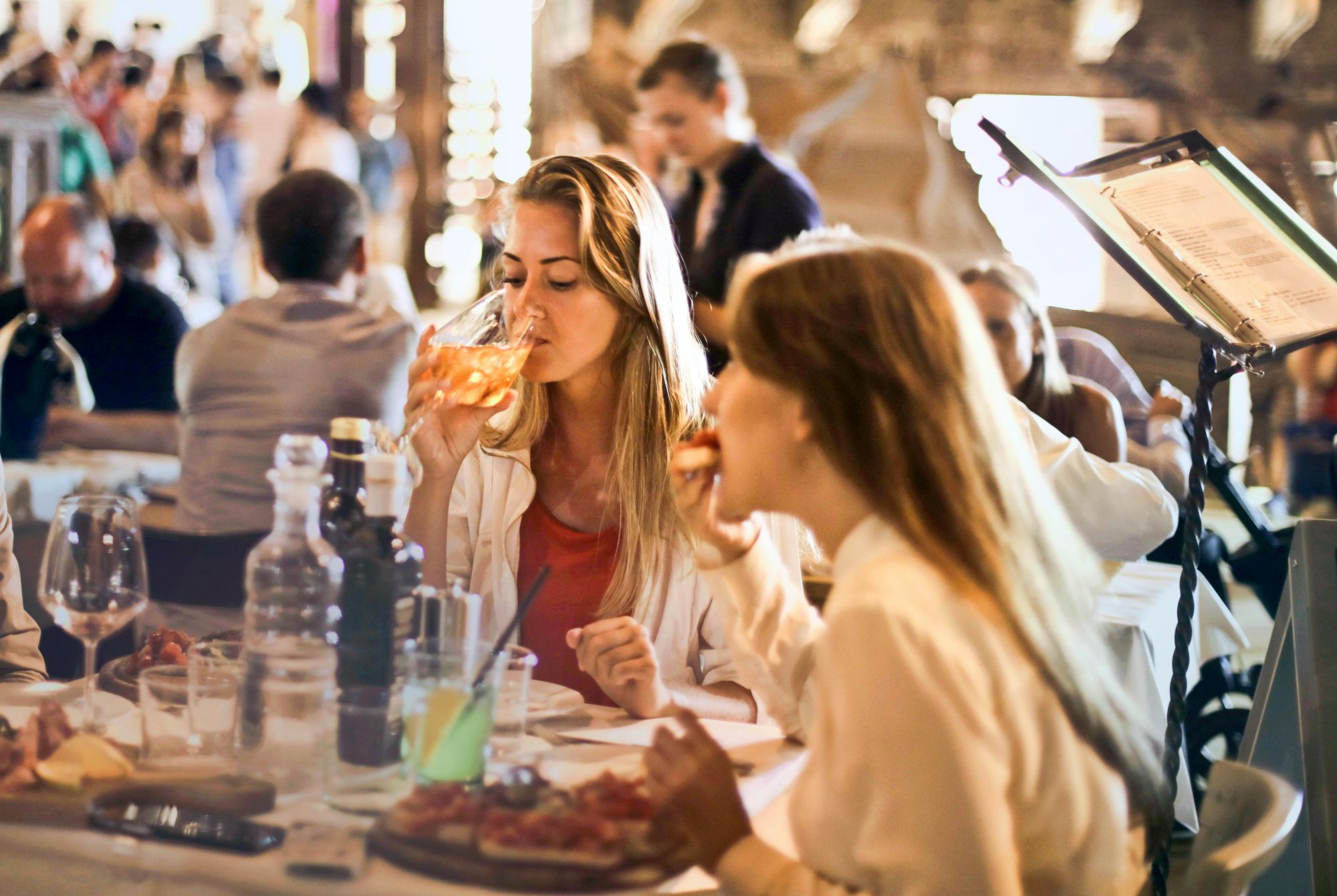 Kobiety pijące drinki w restauracji, w tle goście i podawane jedzenie.